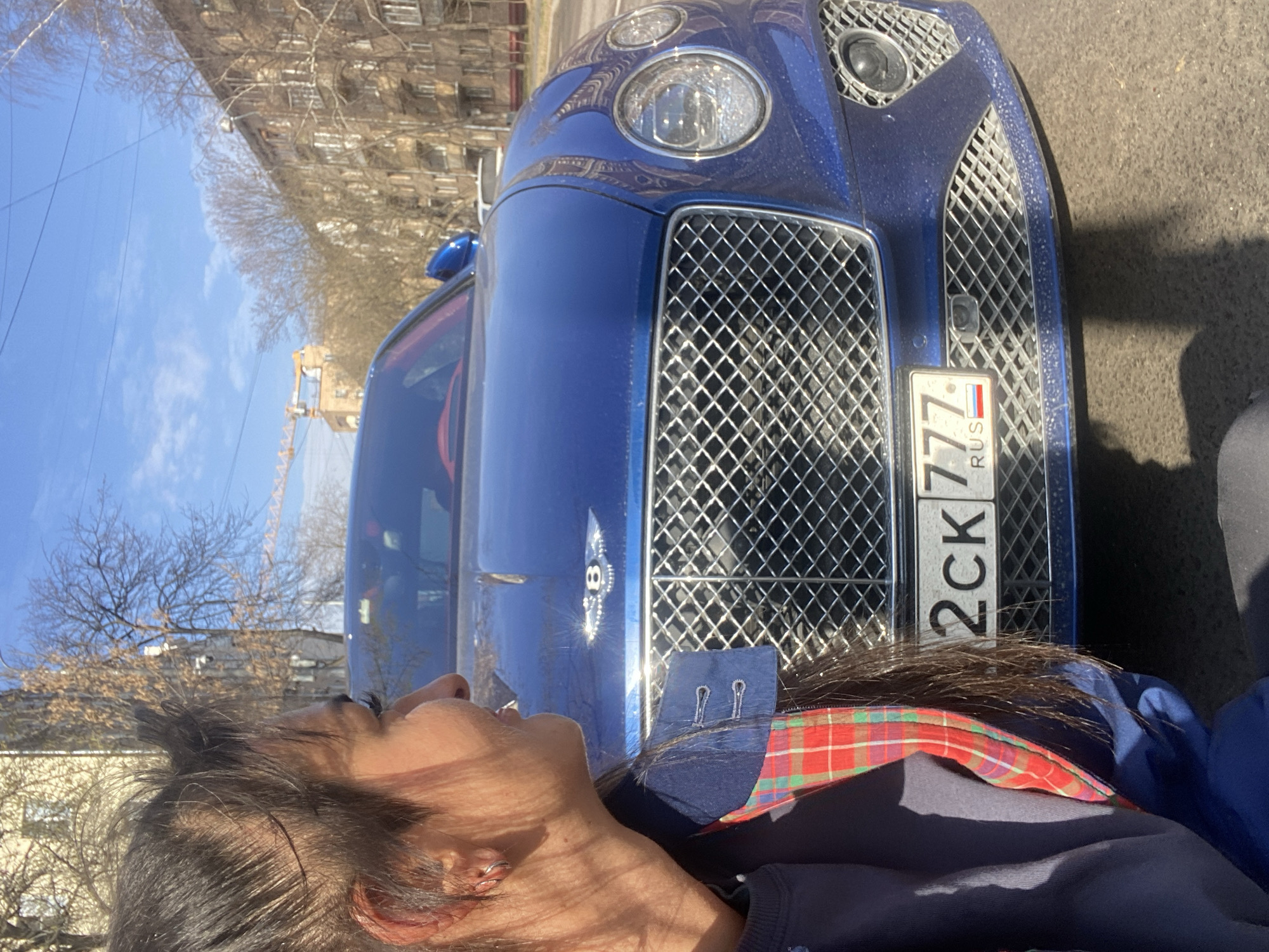 The image shows a person leaning against the hood of a blue Bentley automobile with a Russian license plate "2CK 777 RUS." The background suggests an urban environment, with buildings and bare trees lining the street, consistent with the architecture found in Moscow. The sky is clear and blue, indicating a sunny day.