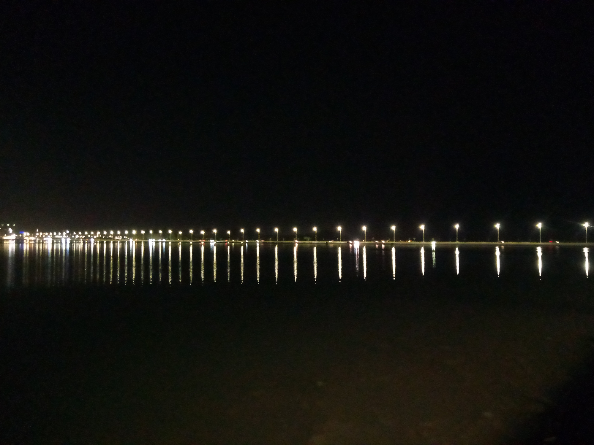 The image depicts a long, straight line of illuminated lampposts stretching across the horizon at night. The lampposts are spaced evenly apart and cast reflections on the still body of water below. The water is dark and reflects the lights, creating a symmetrical pattern. The sky is completely black, indicating nighttime. The scene appears to be a waterfront area or a road alongside a large body of water. This location is likely the Al Khobar Corniche in Al Khobar, Saudi Arabia, a well-known waterfront promenade lined with lampposts and offering views of the sea. There are no people or active events visible in the image, presenting a serene and static nocturnal landscape.