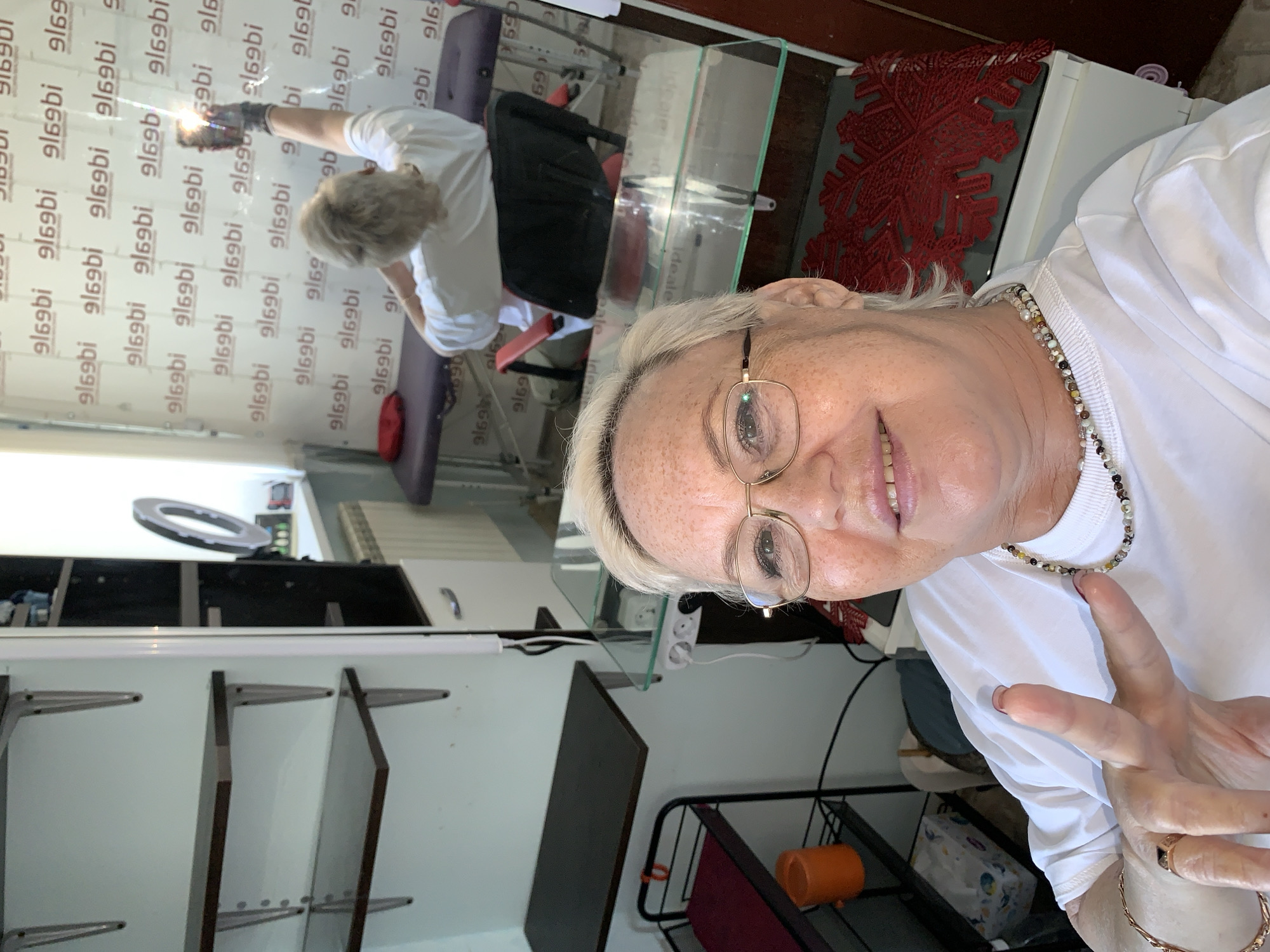 A woman with blonde hair and glasses is taking a selfie in a room that appears to be a beauty salon in Chaikovsky, Russia. She is wearing a white t-shirt and a beaded necklace. Her left hand is raised, making a peace sign. In the background, a woman with blonde hair is visible, sitting on a massage table and holding a smartphone in her raised hand. The room has shelves, a desk with electrical outlets, and a massage table. The walls are decorated with a repeating pattern.