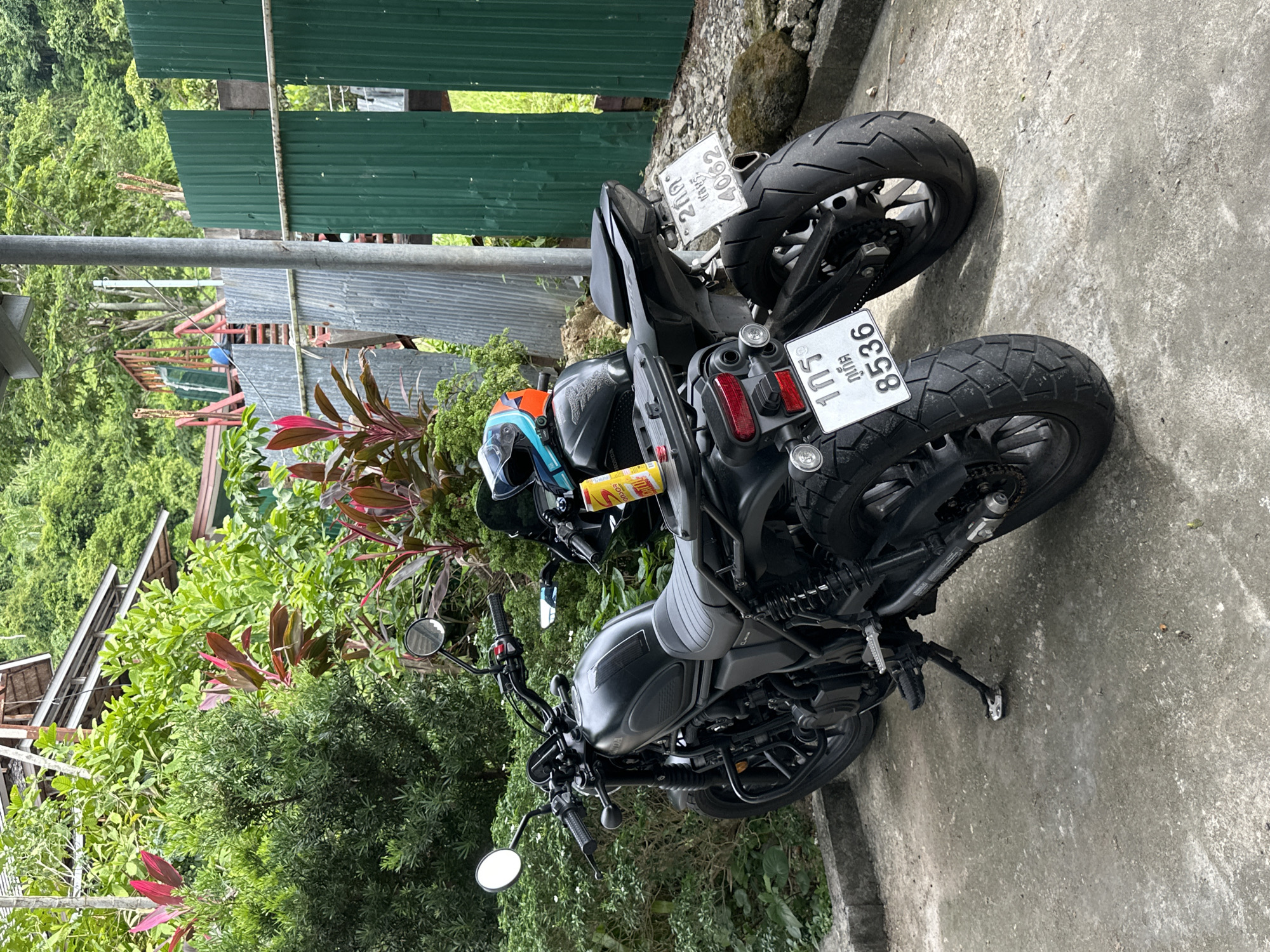 Two motorcycles are parked on a concrete surface next to lush green foliage. The motorcycles are dark in color, with one appearing to be a sportbike and the other a cruiser. Both have visible license plates. The sportbike, closer to the background, has a license plate with the characters "200 4062" and some Thai script. The cruiser, in the foreground, has a license plate that reads "105 8536" also with Thai script. A blue and orange helmet rests on the sportbike. The scene appears to be outdoors in a tropical location, possibly Karon, Thailand, indicated by the dense vegetation and the style of buildings visible in the background, which include corrugated metal roofing. The lighting suggests it is daytime, with soft, diffused light, possibly indicating an overcast sky or a shaded area. The ground is wet, suggesting recent rain. There are no people visible in the image, and the overall mood is peaceful and still, with the focus entirely on the parked motorcycles and their surroundings. A yellow and red packaged item is attached to the sportbike.