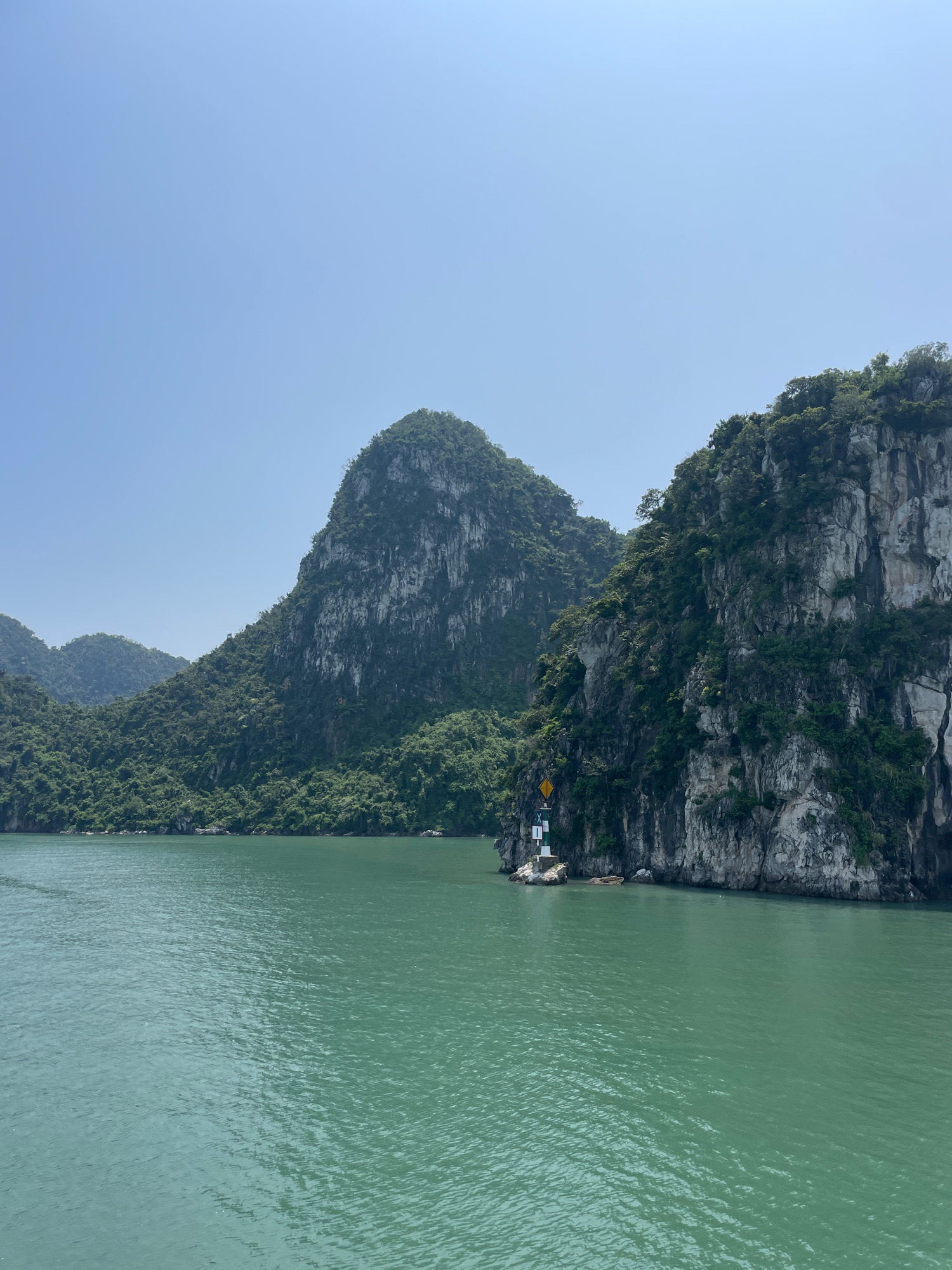 A wide, calm, turquoise body of water occupies the foreground and midground of the image. In the midground, a series of towering, green, vegetated limestone karsts rise from the water. The karsts are steep and rocky, with dense foliage covering their surfaces. A small, white and green lighthouse with a yellow warning sign is situated on a rocky outcrop to the left of the largest karst in the midground. The sky above is clear and blue, with no clouds visible. The scene appears to be a natural landscape, likely a bay or a gulf, possibly located in Vietnam, known for its karst formations.