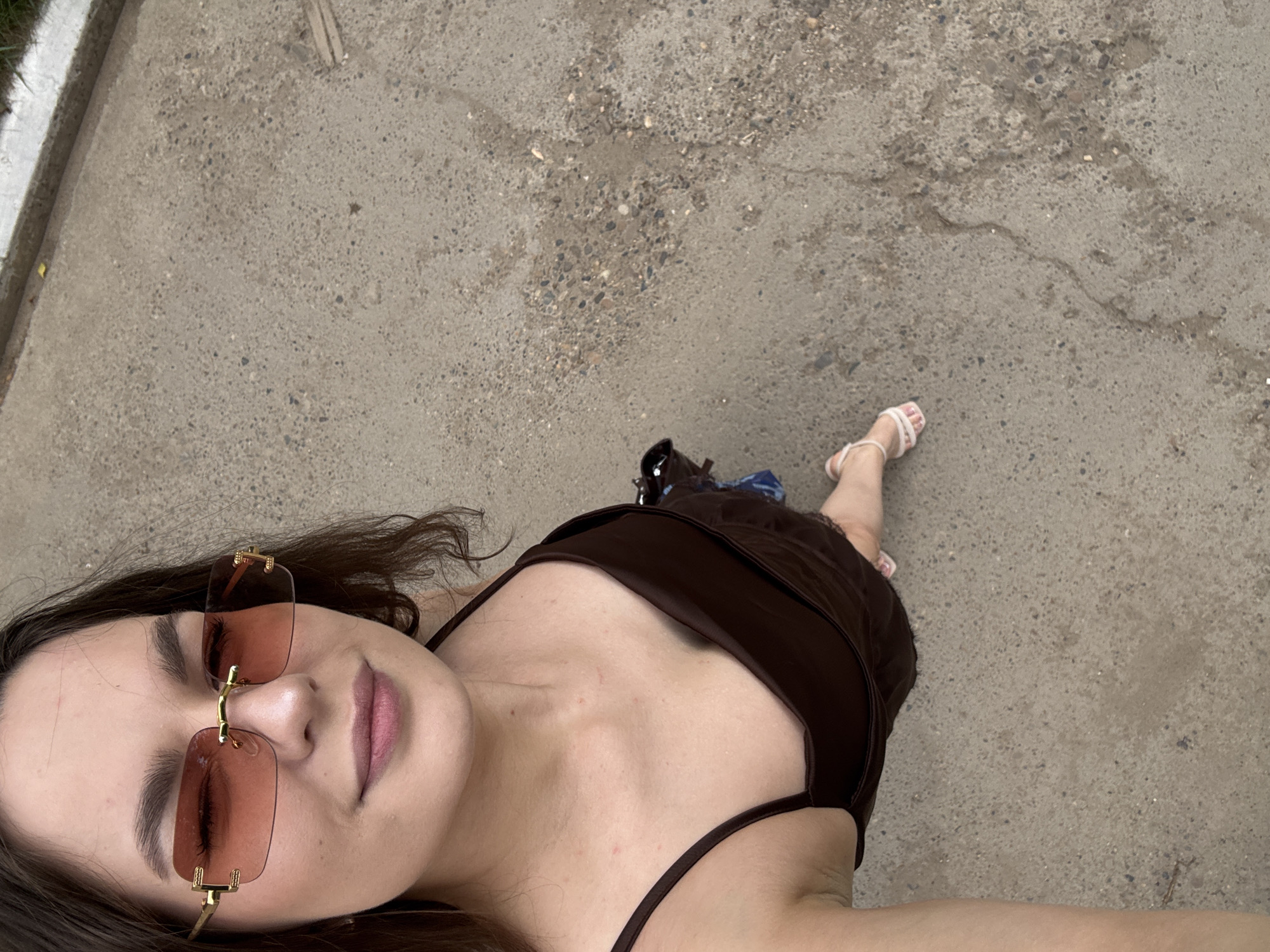 A young woman with dark hair and a brown dress stands on a concrete surface. She is wearing sunglasses with pink lenses and gold frames. Her left arm is extended, holding what appears to be a phone, implying she is taking a selfie. The background is a textured concrete surface with small pebbles and some debris, characteristic of an outdoor urban environment. The lighting suggests it is daytime. The location is likely Chaikovsky, Russia, as per the prompt, though no specific landmarks are visible to confirm this.