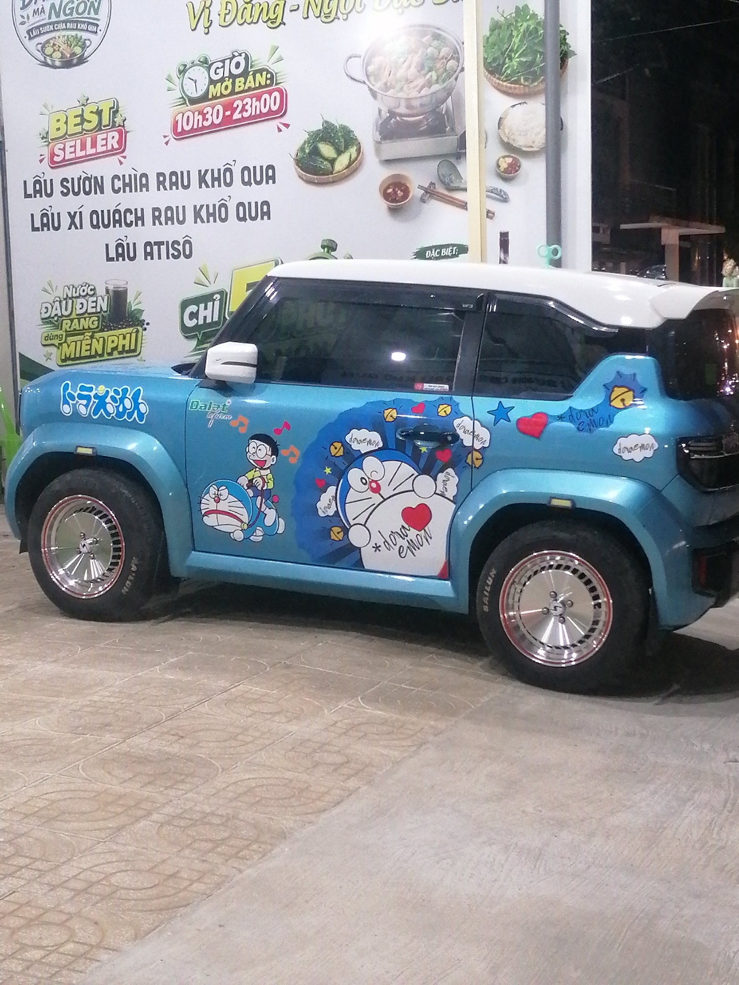 The image shows a blue car decorated with Doraemon stickers parked on a tiled pavement in Da Lat, Vietnam. The car is a small, modern hatchback with a white roof and a spoiler. The stickers depict the cartoon character Doraemon and other related imagery.
Behind the car, a large white sign with Vietnamese text is visible. The sign appears to be an advertisement for a restaurant or food establishment, featuring images of food and promotional messages such as "BEST SELLER" and opening hours. The text on the sign is in Vietnamese, and some phrases are decipherable like "GIỜ MỞ BÁN" (opening hours) and "LẨU SƯỜN CHÌA RAU KHỔ QUA" (rib hot pot with bitter melon vegetable).
The environment suggests an urban setting, likely a street or an outdoor dining area. The lighting indicates it might be daytime, or the area is well-lit. The pavement has a distinctive pattern of circular motifs. The overall impression is a roadside scene in a Vietnamese city, possibly a popular tourist spot given the Doraemon theme.