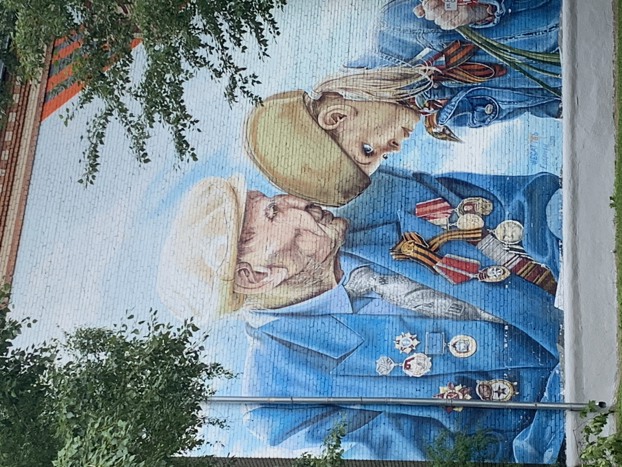 A mural on a brick building depicts an elderly man and a young girl. The man wears a blue jacket adorned with numerous medals and a beige cap. He is leaning down, looking intently at the girl. The girl, also wearing a beige cap and a blue denim jacket, looks up at the man. She holds a bouquet of green stems in her right hand. The background of the mural is a light blue sky with subtle white cloud-like patterns. The mural is situated in an outdoor environment, with green trees partially obscuring the upper portion of the artwork on both sides. A metal pole stands vertically to the left of the man. The lower part of the mural is bordered by a white concrete ledge, with grass visible in the foreground. Faint artist signatures and text are present on the lower right portion of the mural.