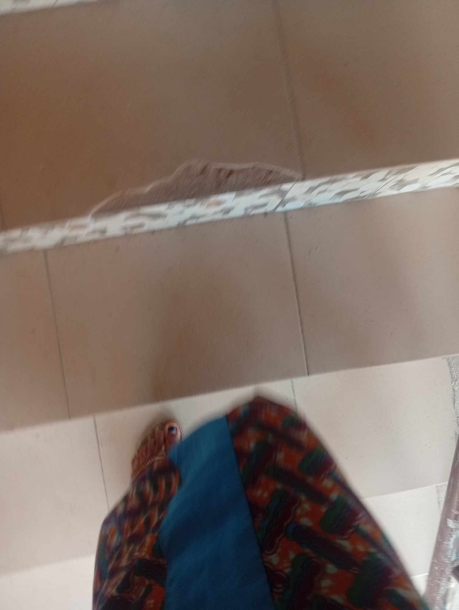 This is a close-up, slightly blurry, low-angle shot taken indoors on a tiled surface, likely stairs or a raised platform. The primary subjects are the lower legs and feet of a person. The person is wearing a colorful, patterned skirt or wrap, with dominant colors of orange, red, purple, and green, and a solid blue garment covering part of the leg. The person is also wearing sandals, one of which is partially visible, revealing a toe.
The tiled surface appears to be a neutral, light brown or beige color, with grout lines visible. There is some damage or wear visible on the edge of the surface in the background, with chunks missing and a rough texture exposed. The lighting suggests it is daytime, but there are no clear indicators of the time of day or weather conditions. The overall impression is of a candid, everyday moment, possibly the person walking or standing on stairs. The blurriness and close-up perspective make it difficult to discern specific emotions or a precise location beyond the tiled environment. No text is visible in the image. The context provided, Jalingo, Nigeria, suggests a potential geographical location for this scene.