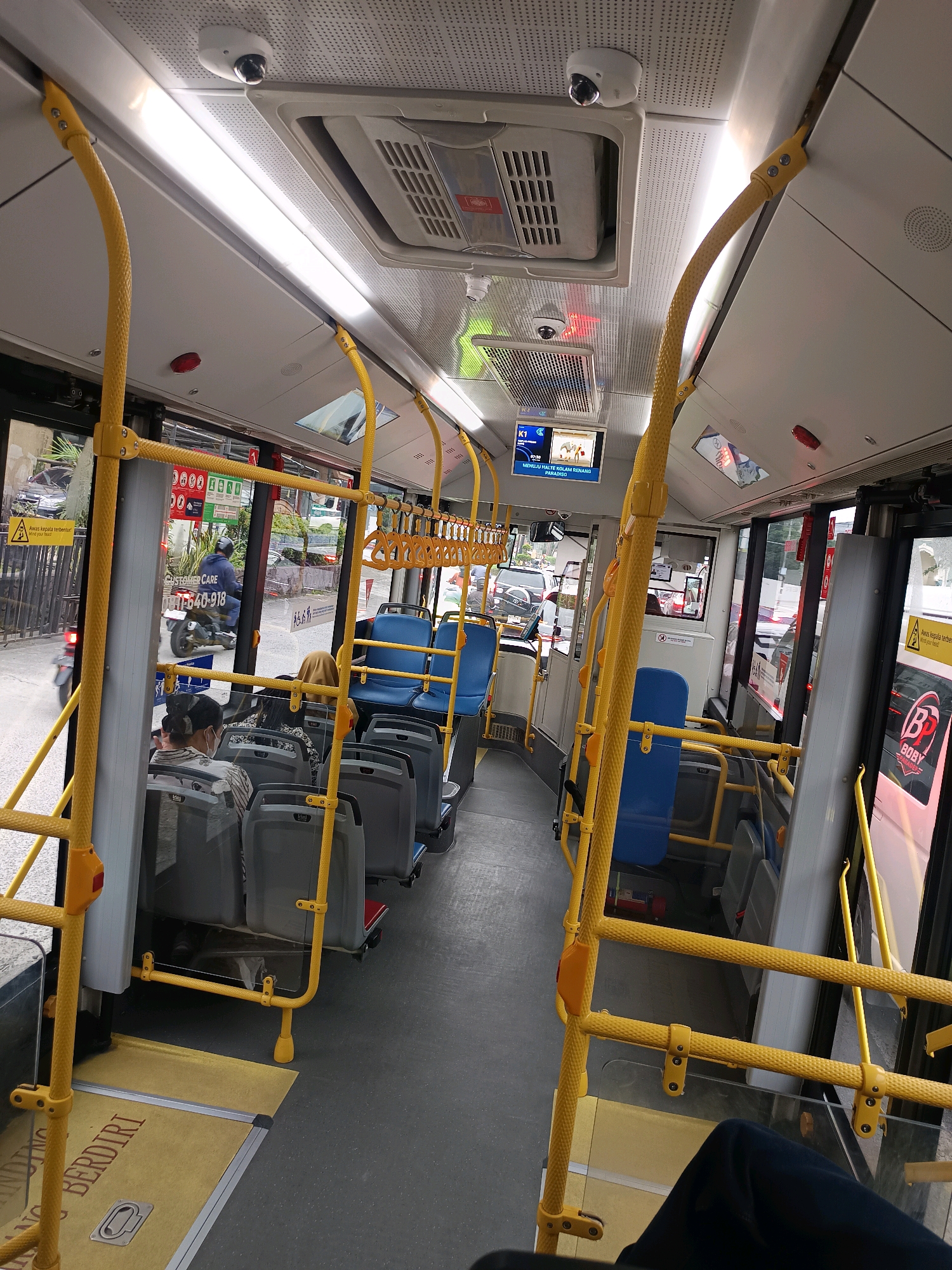 The image captures the interior of a public bus, likely in the City of Medan, Indonesia, as indicated by the text on the yellow floor mats which reads "BERDIRI" (stand) and suggests a transit environment. The bus features rows of blue and gray passenger seats, with prominent yellow safety bars and handrails throughout the aisle and along the ceiling. Several passengers are seated, some wearing face masks. Outside the bus windows, glimpses of urban traffic and pedestrians are visible. Security cameras are mounted on the ceiling. A digital display screen above the seats shows route information. The overall impression is of a functional public transportation vehicle in use.
