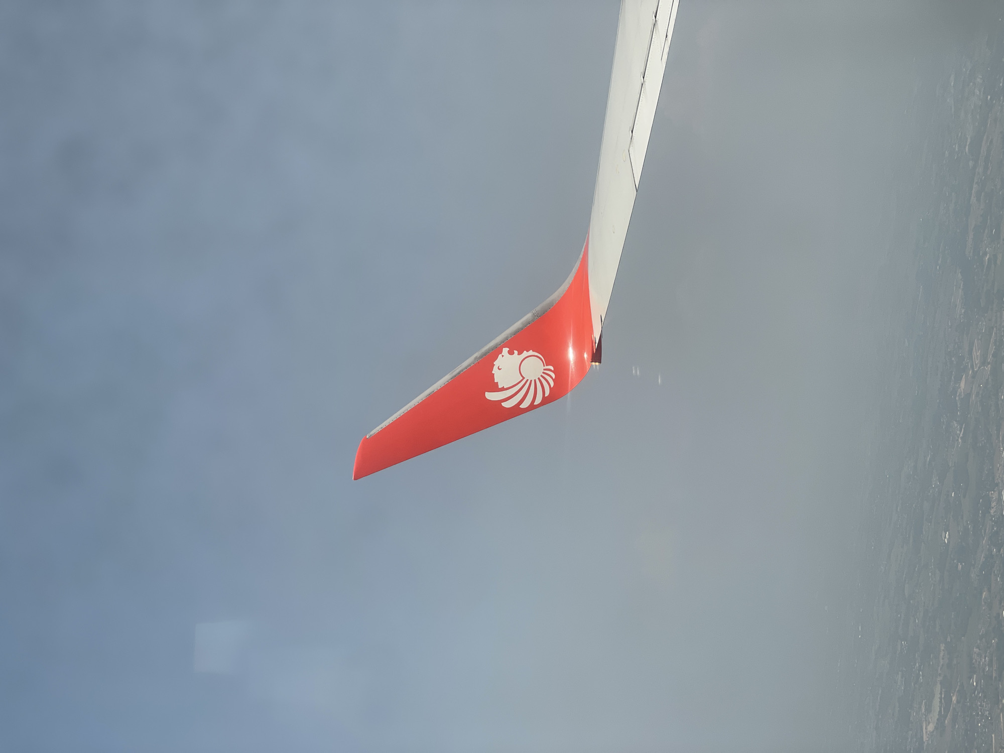 An aerial photograph taken from an airplane window showcases a portion of the aircraft's wing and winglet against a hazy sky. The dominant feature is the distinctive winglet, which is bright red and prominently displays a white, stylized lion's head logo. This emblem is associated with Lion Air Group, suggesting a flight with an airline like Thai Lion Air, fitting the Bangkok, Thailand context.
The main wing, extending upwards and to the right from the winglet, is light grey or white. The background is a soft, uniform greyish-blue, indicating either an overcast sky or significant atmospheric haze. In the distant lower-right, a faint, blurred outline of a landscape or urban area can be discerned beneath the atmospheric conditions, consistent with looking down at a city like Bangkok from altitude. The image captures a serene moment during daytime flight. No people are visible.