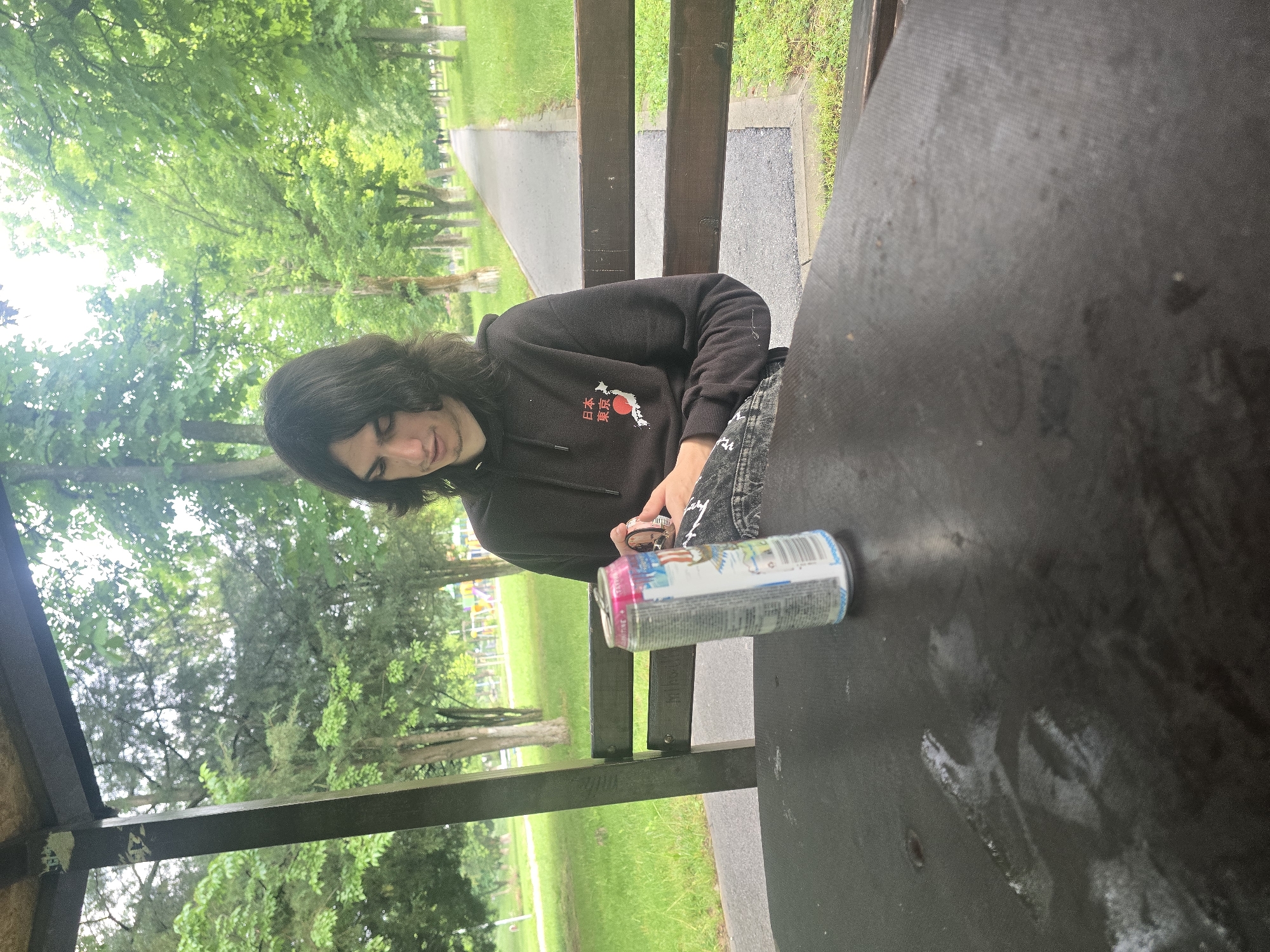 The image shows a young person seated at a dark-colored table, likely under a shelter or gazebo in a park. The photo is rotated, making the person appear to be on their side.
The main subject is a person with long, dark hair, wearing a black hooded sweatshirt and patterned jeans. On the front of the hoodie, there is a graphic featuring a red circle over a white outline of the map of Japan, accompanied by Japanese characters that translate to "Japan Tokyo." The person is looking downwards, with their hands engaged in an activity, possibly manipulating or opening a beverage can.
A colorful can, predominantly pink, blue, white, and silver, lies on the table in front of the person. Partial text "ULTRA" is visible on the can, suggesting it might be an energy drink. The table itself is dark and shows some lighter scuff marks or chalk residue.
The background reveals a verdant outdoor setting, characteristic of a park. Numerous trees with lush green foliage and green grass are visible under natural daylight, suggesting it is daytime. A paved pathway winds through the park in the distance. Wooden beams of the shelter structure are also visible, framing the scene. The overall impression is a casual moment in a public green space.