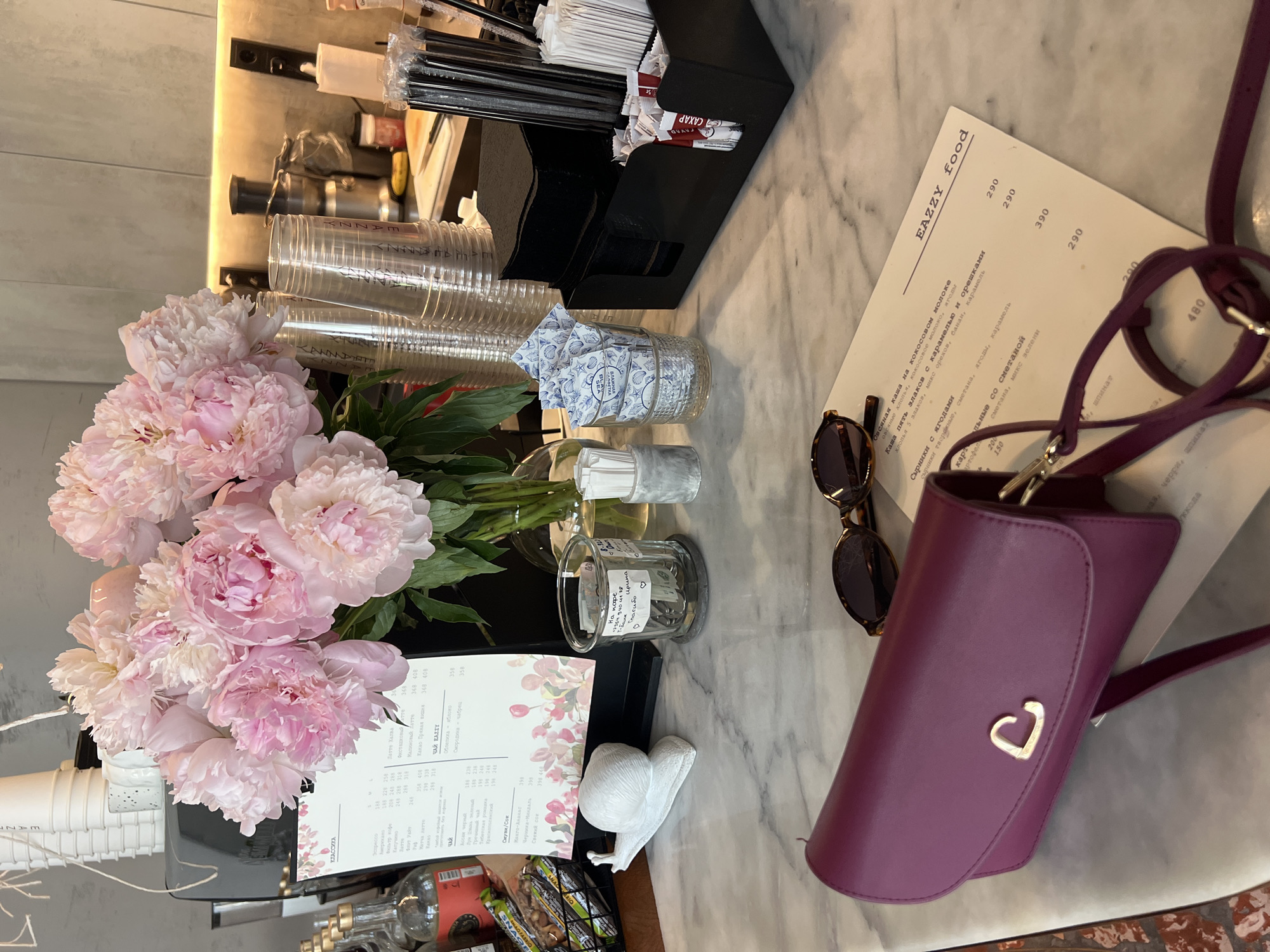This is a top-down, eye-level shot of a marble-topped surface in what appears to be a cafe or restaurant. The scene is artfully arranged, suggesting a focus on aesthetics and perhaps the cafe's offerings.
In the foreground, a bouquet of large, fluffy pink peonies is the dominant subject, their soft petals filling a significant portion of the left side of the frame. To their right, a small, clear glass jar holds what look like sugar packets, some with a shell pattern and text in Russian. Next to it, another glass container holds packets of sugar labeled "CAXAP" and "EAZZY food" in English.
Further to the right, a stylish purple clutch purse with a gold heart detail lies open, revealing a strap. A pair of dark sunglasses rests on top of a menu. The menu, also featuring Russian text, has a title "EAZZY food" and lists various items like "Овсяная каша на кокосовом молоке" (Oatmeal on coconut milk), "Каша пять злаков с карамелью и орешками" (Five-grain porridge with caramel and nuts), and "ЧАЙ EAZZY" (EAZZY Tea). Prices are listed alongside the items.
In the background, a black organizer holds an assortment of items, including black straws and white paper packets. Clear plastic cups are stacked to the left of the flowers. The overall lighting suggests it could be daytime, possibly late morning or early afternoon. The marble surface and the elegant arrangement of items imply a sophisticated and well-maintained establishment, likely in an urban or resort setting, given the context of Sochi. The overall mood is calm and inviting, with the flowers adding a touch of natural beauty.