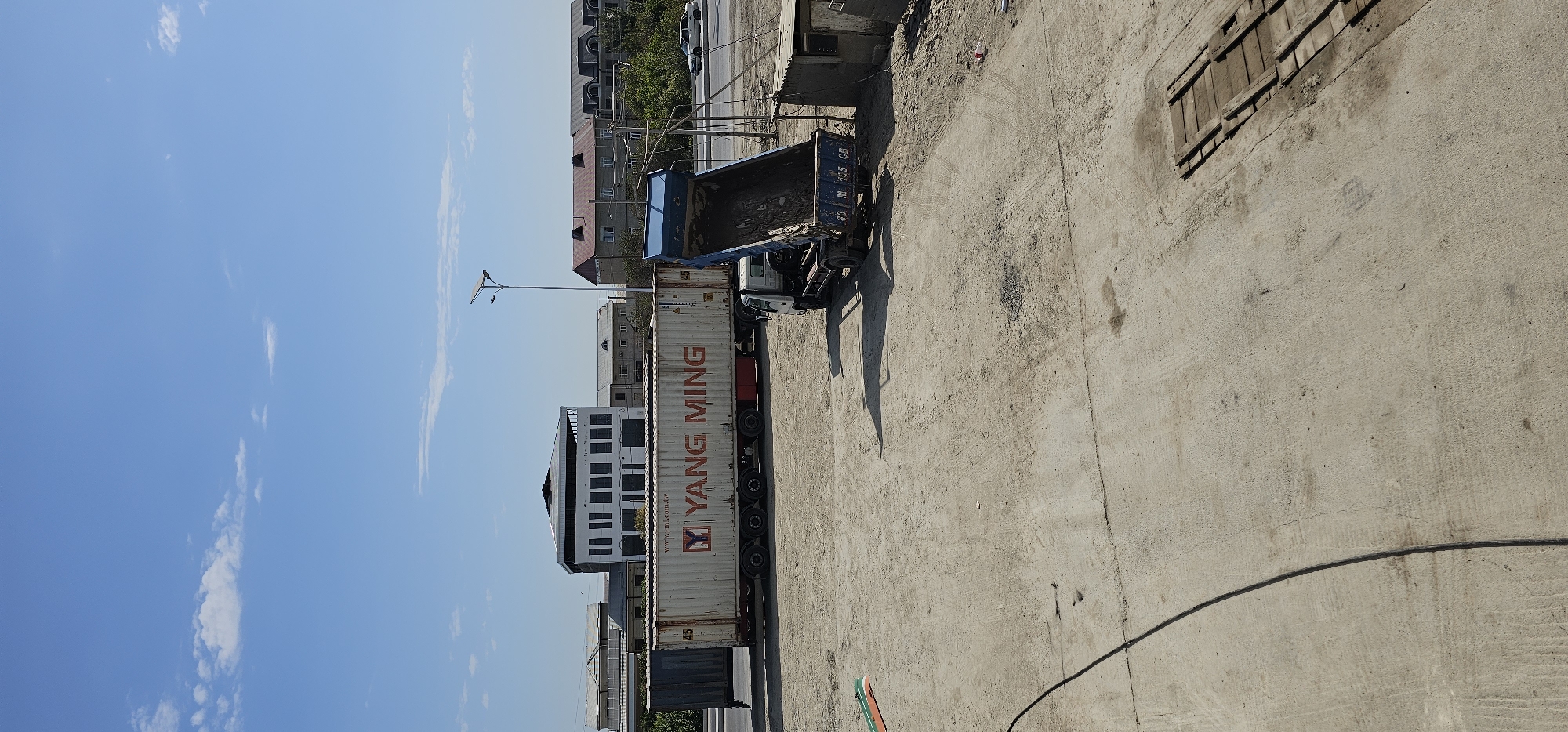 An outdoor, daylight scene in Sheyhancha, Uzbekistan, captures a logistics and construction area. Dominating the mid-ground is a large white shipping container bearing the red "YANG MING" logo, mounted on a white semi-trailer truck. To the right, a blue dump truck is positioned with its bed raised, appearing to be in the process of dumping a load of dark material. In the background, several multi-story buildings with distinct architectural styles line a road where a white car is visible. A solar-powered street lamp stands tall on the right side of the frame. The ground is a mixture of compacted dirt and concrete, with tire tracks suggesting vehicle movement. A small, dilapidated shed-like structure is situated near the dump truck.