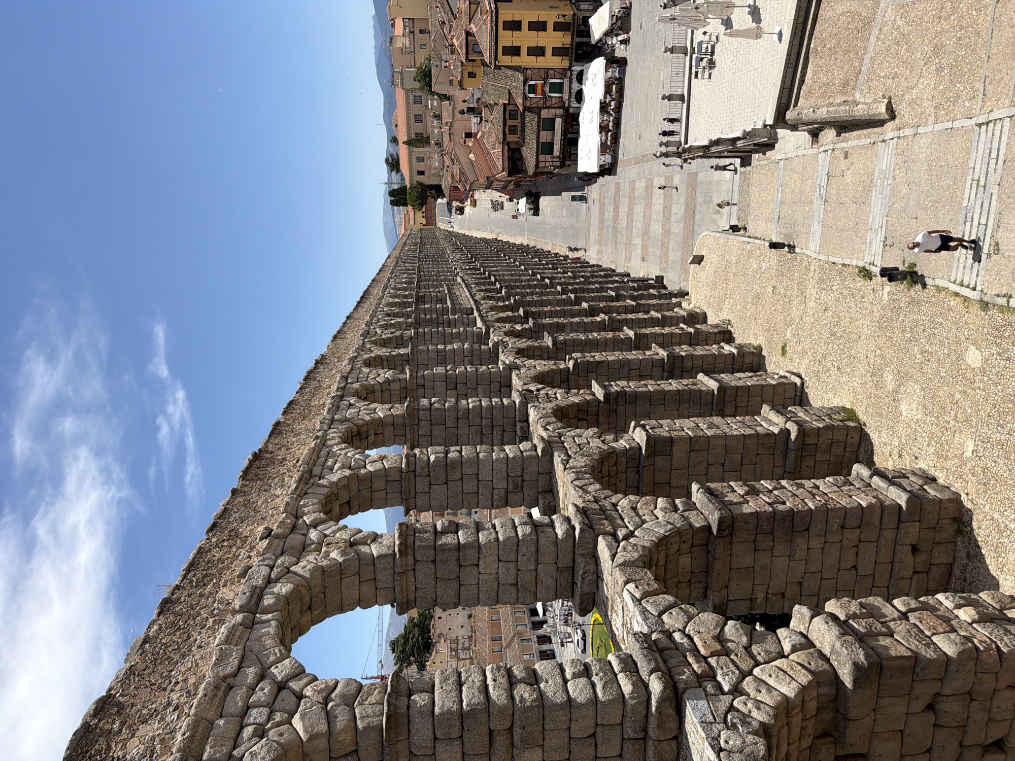 The image shows the Aqueduct of Segovia, Spain, a well-preserved Roman aqueduct that is a UNESCO World Heritage site. The aqueduct is constructed from large granite blocks and features a double tier of arches. In the foreground, the massive stone structure dominates the left side of the frame, stretching into the distance. The right side of the image reveals a city street lined with buildings, some with traditional tiled roofs and vibrant facades. People are visible walking along the street and on stairs leading up to a plaza area. The sky is clear and blue with scattered white clouds. The overall scene depicts a sunny day in Segovia, with the ancient aqueduct serving as a striking backdrop to the modern city life.