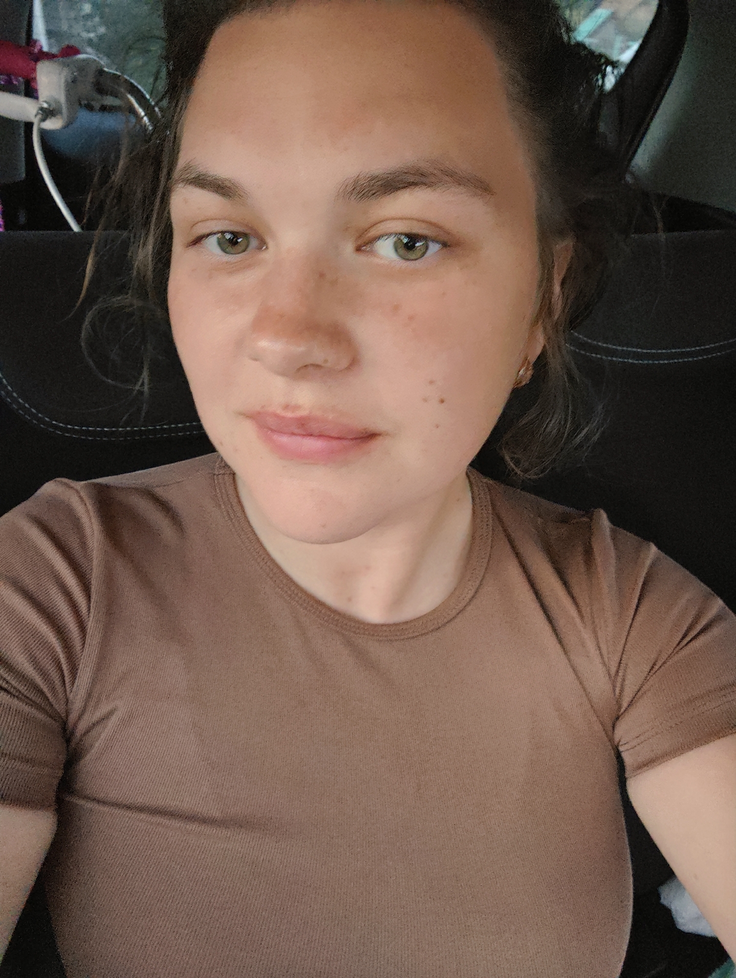 A woman is positioned in the driver's seat of a vehicle. The woman has fair skin, freckles, and dark hair. She is wearing a brown t-shirt. The background is indistinct, but suggests the interior of a car, including the back seat with a USB charging port and a small cable. The environment and the objects visible do not provide any information regarding the location in the city of Уральское, Russia.