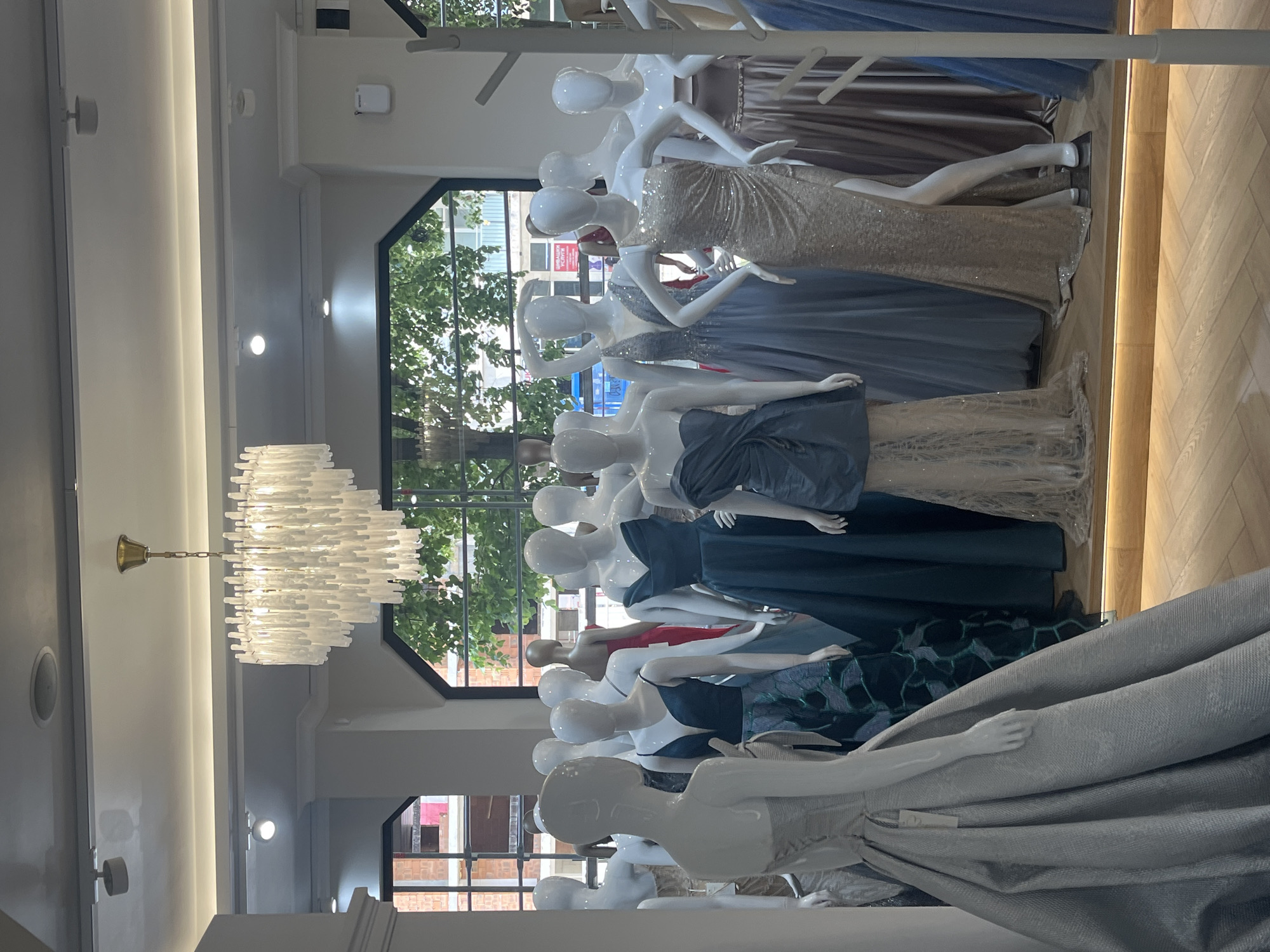 The image shows a fashion store in what appears to be Blagoevgrad, Bulgaria. The interior features a series of mannequins displaying various dresses. The dresses range in color and style, from shimmering gold to deeper blues and patterned designs. The mannequins are arranged in a line, facing the window. A chandelier hangs from the ceiling, adding to the store's ambiance. Outside the window, trees and buildings are visible, suggesting a city setting.