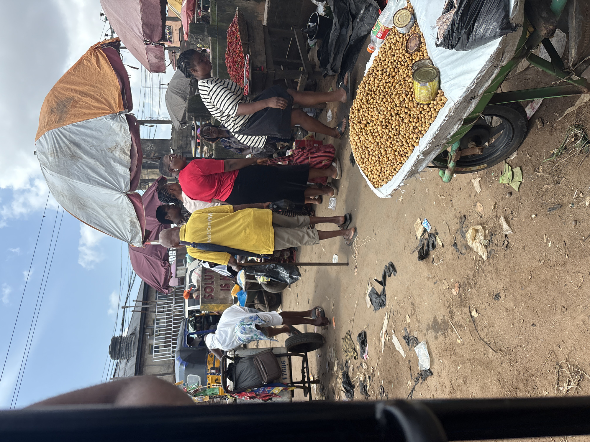 The image depicts an outdoor market scene in Fegge, Nigeria. In the foreground, a large cart filled with dried, light brown legumes or nuts is prominently displayed. Several cans, including an open yellow one, sit atop the legumes. To the right of the cart, a woman in a black and white striped shirt and a black skirt is leaning over, examining goods at a stall. Behind her, another woman is visible. Further back, several other individuals are present, some standing under large, colorful umbrellas. One man in a bright yellow shirt with "HealthCarePH" visible on the back stands with a stick. To the left, a cart with a white sheet draped over it is being tended to by a man in white attire. The ground is dirt and littered with various pieces of trash, including plastic bags and paper. In the background, buildings and a water tank can be seen. The sky is partly cloudy.