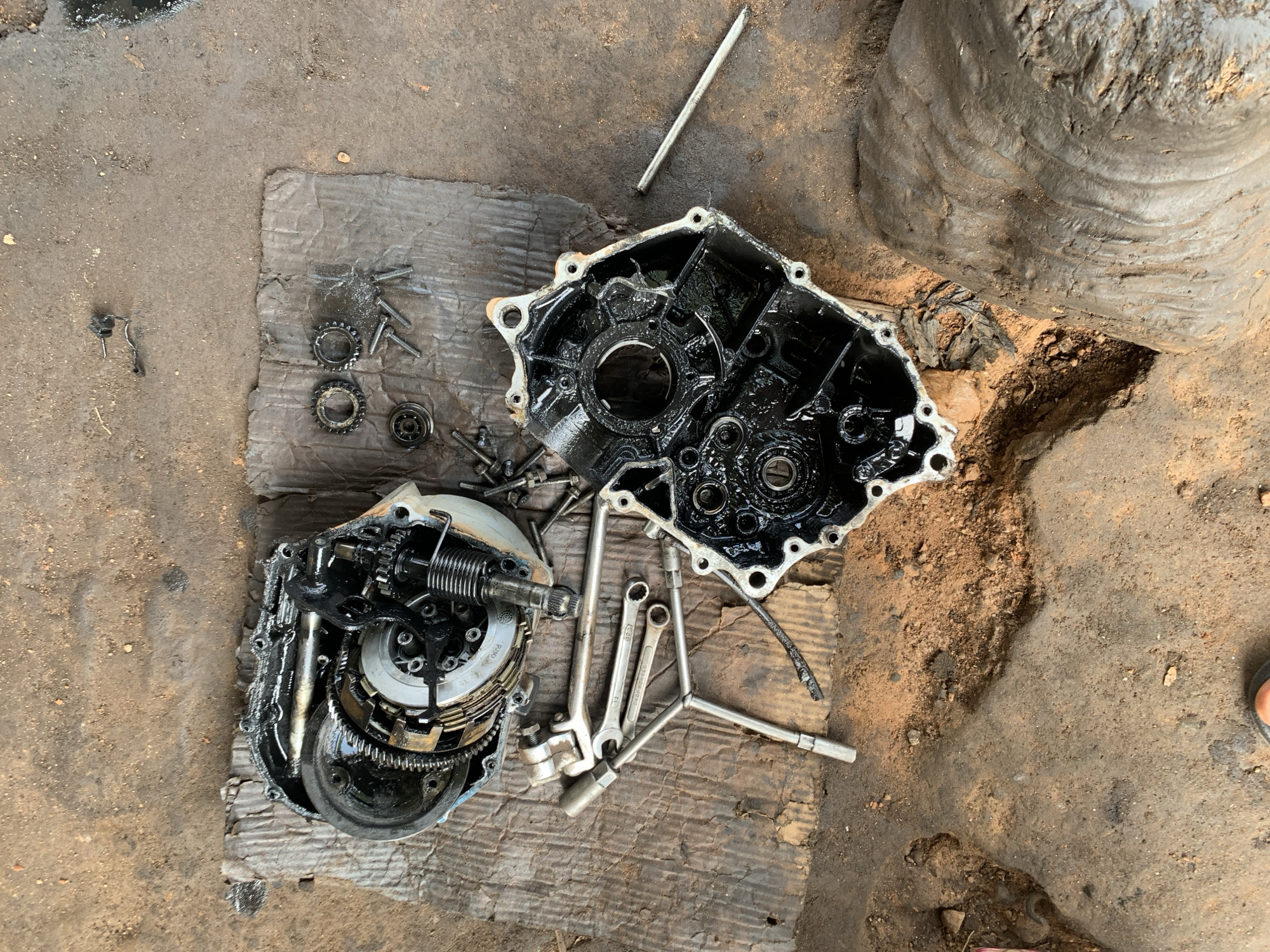 This overhead shot captures a motorcycle engine's disassembled parts laid out on a piece of cardboard on a dirt ground, suggesting an outdoor repair setting. The primary objects are the engine's crankcase, a clutch assembly, and various tools, including wrenches and a socket. The engine parts are visibly greasy, indicating recent disassembly or maintenance. A weathered, grey, cylindrical object, possibly a large tire or a part of a structure, is visible in the background, contributing to the rustic feel of the scene.
The image seems to be taken during daylight, with natural lighting illuminating the workspace. The ground is a mix of dirt and sand, with some scattered debris and oily patches, typical of a workshop or repair area in a place like Kabba, Nigeria. The overall atmosphere is one of practical work, with no discernible emotional cues. The focus is on the mechanical components and the tools used to service them. There is no visible text in the image.