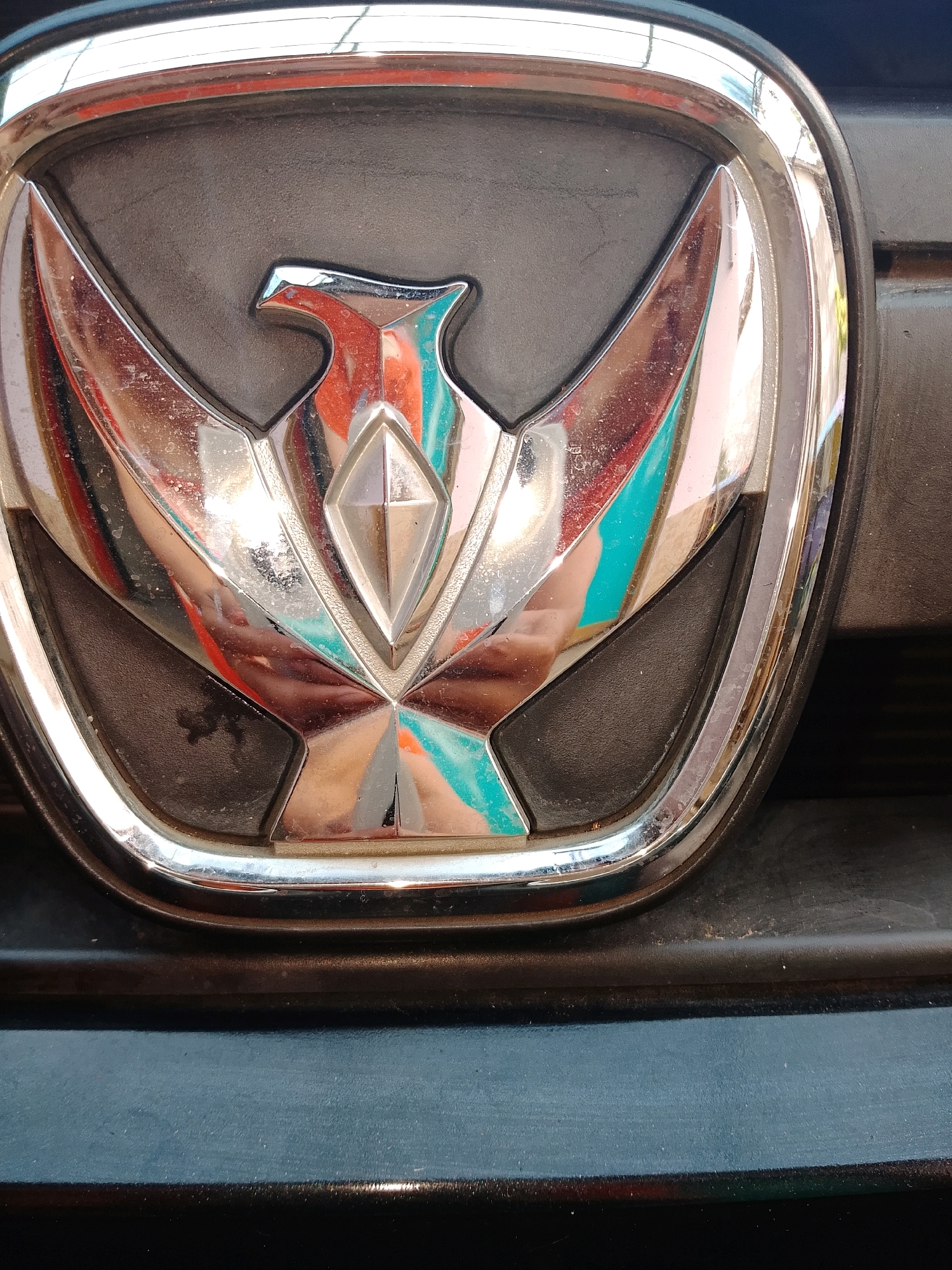A close-up, eye-level, medium shot shows a chrome and black car emblem. The emblem features a stylized bird with outstretched wings, rendered in chrome with red and teal accents. The bird's head is to the left of the frame and its body is in the center, with a diamond shape at its chest. The emblem is set against a black background and is framed by a chrome border. The car is dark-colored and appears to be parked outdoors. The lighting is bright and reflects off the chrome surface. The image was taken in Gebang, Indonesia.