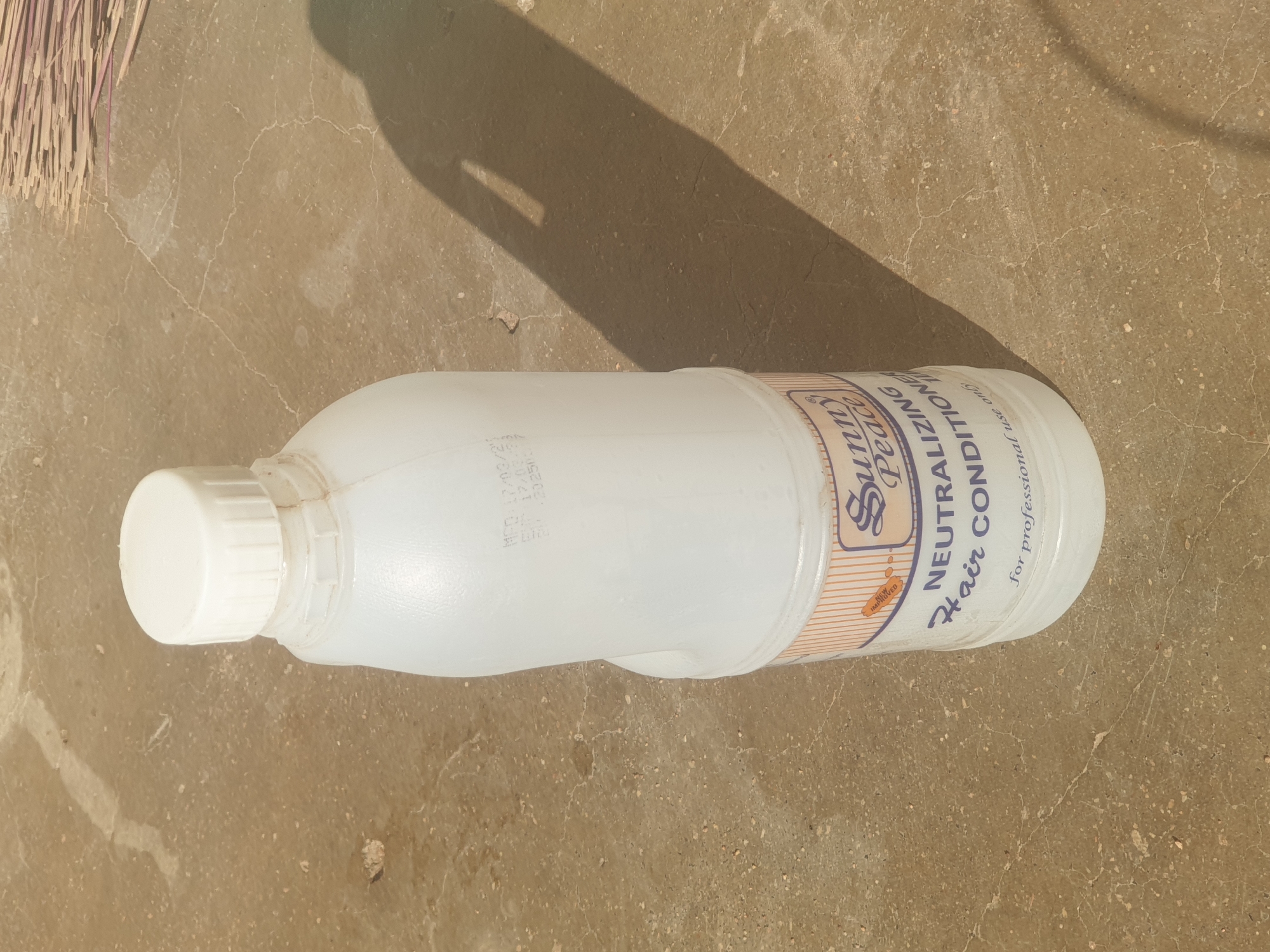 A white, opaque plastic bottle, with a white ribbed screw-cap, is positioned upright on a light brown concrete surface. The bottle features a label with a striped orange and white background, displaying the brand "Sunny Peace" and the product "NEUTRALIZING Hair CONDITIONER," along with the phrase "for professional use only." A small oval graphic with "IMPROVED" is visible above the brand name. Embossed text on the upper part of the bottle reads "MFD: 17/03/21," "EXP: 17/03/23," and "BPL 03250."
The concrete ground is textured with small aggregate and crisscrossed by fine cracks. Strong natural light, likely from the upper right, casts a distinct, elongated dark shadow of the bottle, including the outline of a handle-like indentation, across the concrete to the left. In the upper right background, a collection of thin, light-colored, possibly dried, sticks or reeds is partially visible. No individuals or active interactions are present in the scene. The environment suggests an outdoor setting.