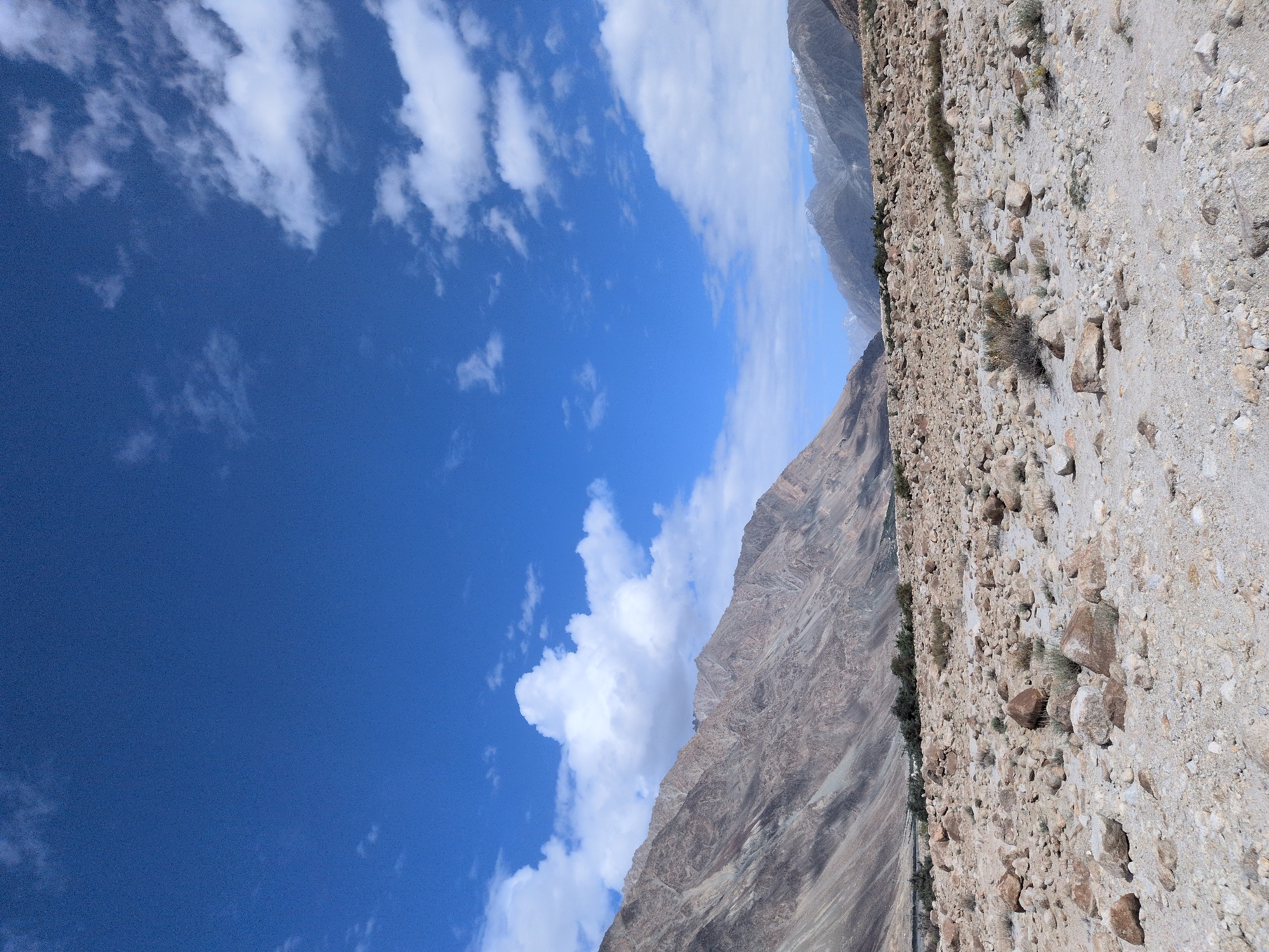 The image depicts a mountainous, arid landscape with a rocky foreground. The sky is a clear blue, dotted with white cumulus clouds. In the background, large, rugged mountains dominate the horizon, their slopes displaying various shades of brown and gray. Some of the peaks in the distance appear to be snow-capped. The ground in the foreground is covered with numerous stones and pebbles of varying sizes, with sparse, dry vegetation growing between them. The overall impression is one of a remote and harsh natural environment. The scene is devoid of any human presence or activity. The location is identified as Sasoma, India.