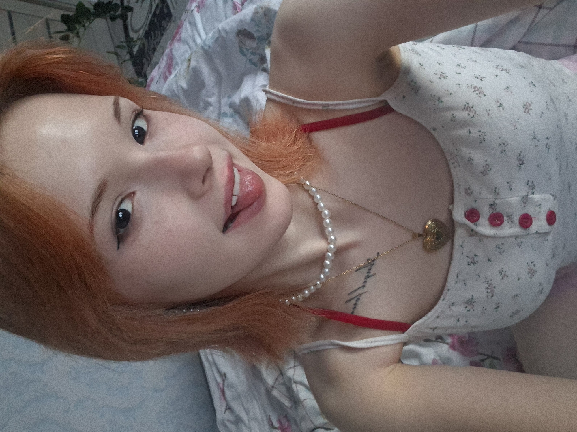 A young woman with reddish-orange hair and fair skin, adorned with light freckles, lies casually, seemingly taking a selfie. She is looking directly at the camera with a playful expression, her tongue slightly out. She wears a light-colored top with a delicate floral pattern and red decorative buttons on a pocket-like detail. A red bra strap is visible beneath her top. Around her neck, she wears a pearl necklace and a gold chain with a small heart-shaped locket. A script tattoo is visible on her upper chest/collarbone area, which, upon closer inspection, appears to read "Туризм" (Tourism) in Cyrillic.
The setting appears to be indoors, likely a bedroom, with the woman resting on a bed covered with patterned sheets, featuring both floral and geometric designs. In the blurred background, green foliage from a plant and possibly another patterned textile or wall hanging can be seen. The lighting suggests daytime, with a soft, even glow. The overall mood is relaxed and cheerful. The context provided indicates the location is Chaikovsky, Russia, which aligns with the visible Cyrillic text.
