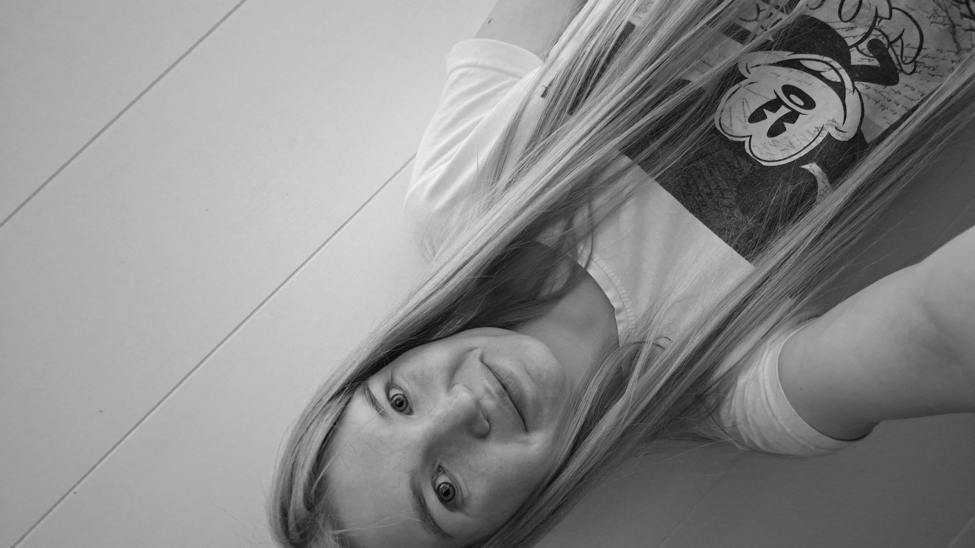 A black and white, close-up, selfie-style portrait of a young woman with long, blonde hair is shown. She is wearing a t-shirt with a graphic of Mickey Mouse on it. The background appears to be a wall with horizontal lines, possibly paneling or siding. The woman is looking towards the camera with a slight smile. The lighting is soft, and the image is slightly grainy. The location is not discernible from the image itself, and therefore, cannot be specified as Большой Букор, Russia.