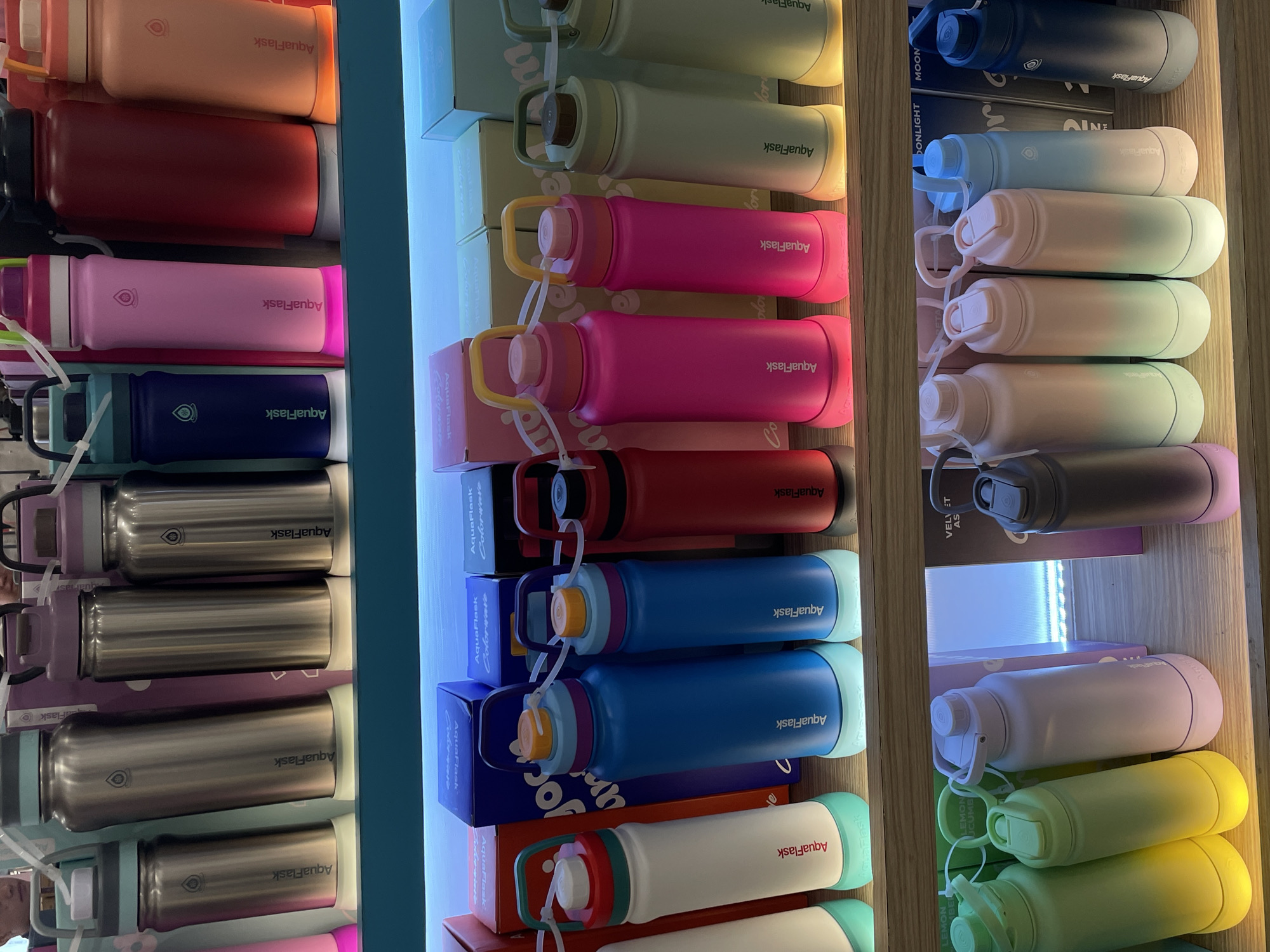 The image displays a retail display of AquaFlask brand water bottles in various colors and sizes, arranged on wooden shelves. The bottles are primarily made of stainless steel and feature a variety of lid designs, including screw-top and flip-top. Some bottles have colorful ombre designs, while others are solid colored. The display is lit from below, creating a glow around the base of the bottles. Behind the bottles, boxes with the AquaFlask logo and product names are visible. The setting appears to be a retail store, likely in Davao City, Philippines, given the context. No people are explicitly visible in the image, but the arrangement suggests a commercial environment.