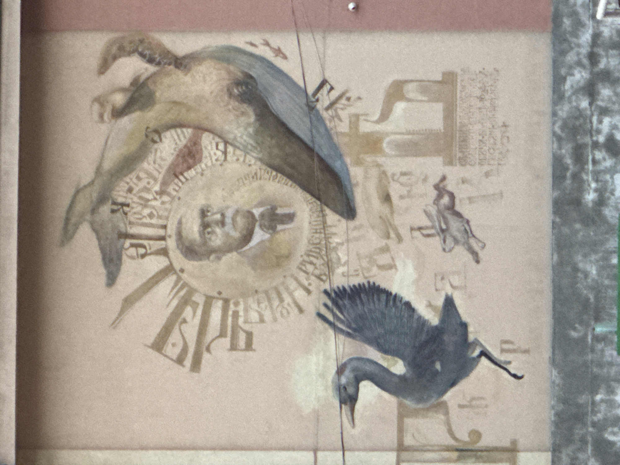 This image depicts a detailed mural painted on the exterior wall of a building in Plovdiv, Bulgaria. The central element of the mural is a sepia-toned bust of a man with a mustache and glasses, wearing a suit jacket and a bow tie, identifiable as the Bulgarian writer Ivan Vazov. He is positioned within a circular motif resembling a sunburst or clock face, surrounded by faded, stylized letters and symbols, predominantly in Cyrillic script.
To the right of the central figure, a large aquatic creature, resembling a whale, is depicted swimming upwards. It features a mottled greenish-brown dorsal side and a lighter blue-grey ventral side, with articulated fins. Below and to the left of the central figure, a large, dark grey bird, appearing to be a crane or heron, is shown mid-motion with its wings partially spread and one leg raised, suggesting either landing or taking flight.
In the background, numerous faded, light brown abstract shapes, architectural outlines, and letters are scattered across the wall. A small, light-colored animal, resembling a rabbit or hare, is depicted running or leaping between the crane and the whale-like creature. At the bottom right, a rectangular block of faint, unreadable Cyrillic text is visible, possibly historical or biographical information.
Two thin, dark electrical wires traverse horizontally across the mid-section of the mural. The bottom portion of the image shows a grey, textured concrete ledge or wall, possibly the top of a lower structure or railing. The far right edge of the frame reveals a vertical section of a solid reddish-brown wall. The entire mural is rendered on a light tan or beige background, and the image exhibits slight graininess.