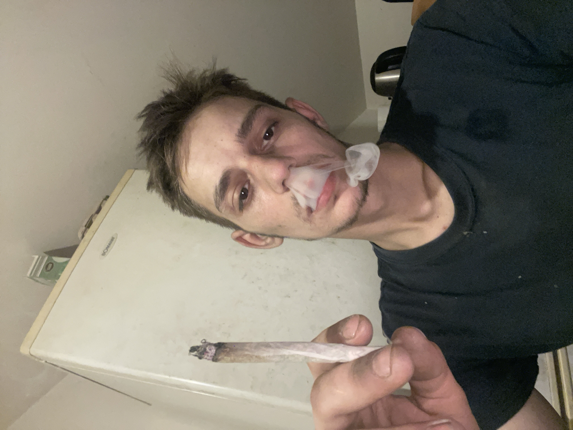 A young adult male with disheveled dark hair is holding a lit cigarette in his left hand and exhaling smoke through his nose and mouth, creating a visible cloud. He is wearing a dark t-shirt. The background appears to be an indoor setting with a white refrigerator on the left and a dark appliance, possibly a kettle, on the right. The lighting suggests it is nighttime or a dimly lit indoor space.