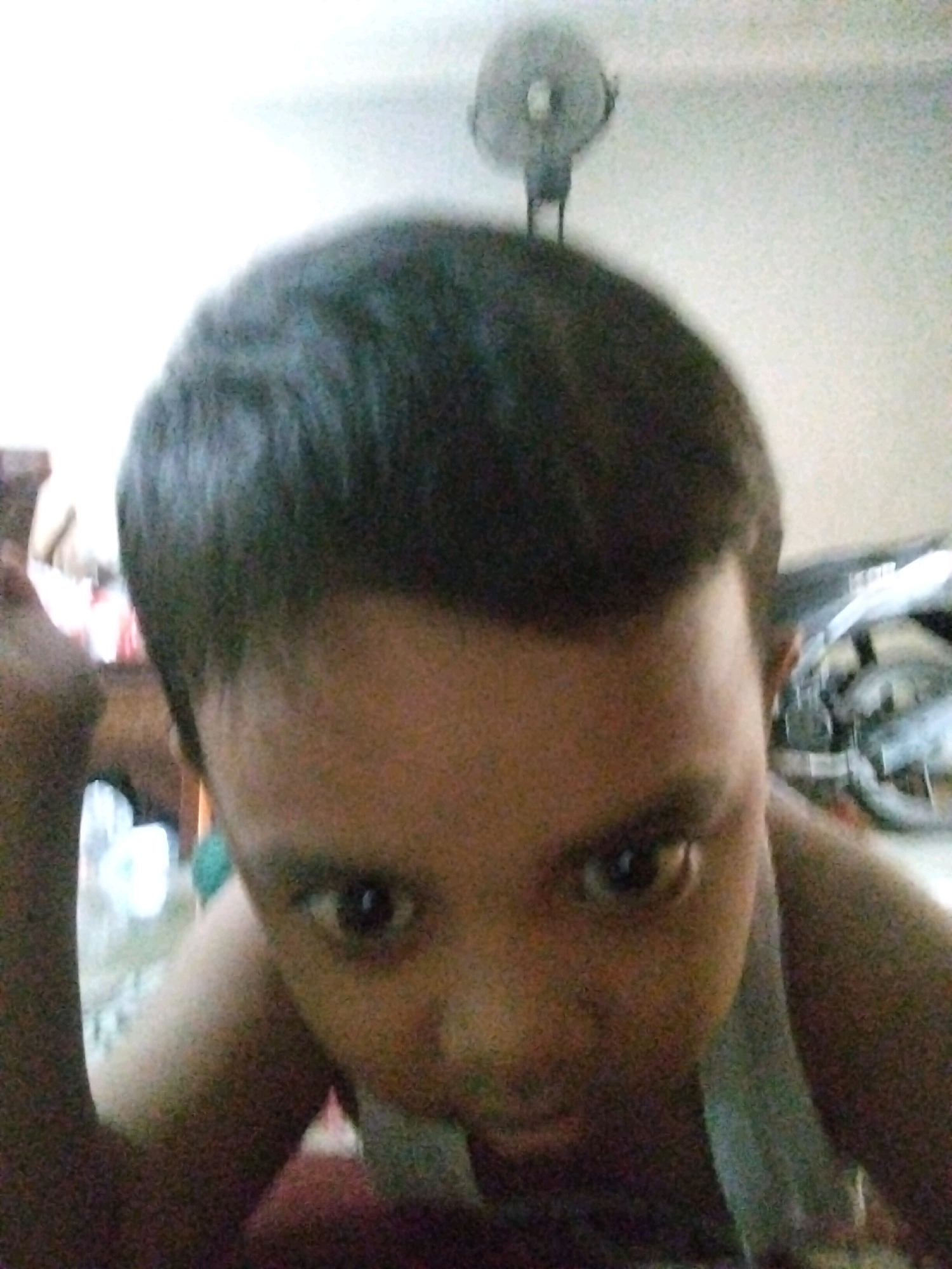 The image is a low-resolution, blurry selfie taken indoors. A young boy with dark hair is the primary subject, with his face filling most of the frame. He is looking directly at the camera, with his head tilted and resting on his left arm, which is bent. His right arm is also visible, extended upwards and partially out of frame. A ceiling fan is visible in the background, out of focus. The background also shows some indistinct objects, including what appears to be furniture and possibly a rolled-up mat or fabric. The lighting is dim, with a bright, blown-out area in the upper left corner. No specific details about the location within Pepelegi, Indonesia, can be discerned from the image.