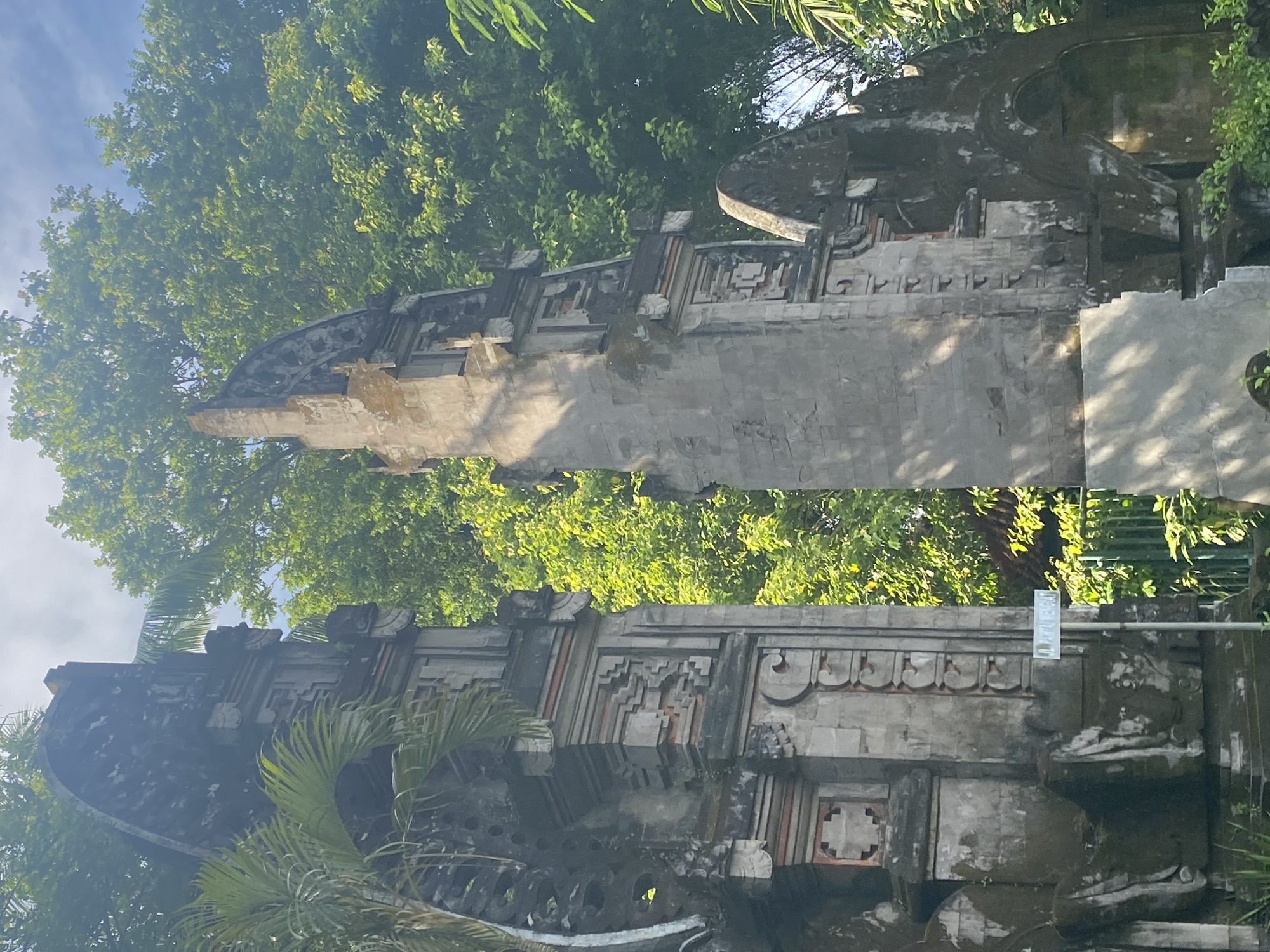 The image depicts a traditional Balinese split gate, likely a "Candi Bentar," against a backdrop of lush green trees in Nusa Dua, Indonesia. The gate is constructed of grey stone, featuring intricate carvings and a weathered appearance. It is composed of two symmetrical structures, separated by a central gap. The structure shows damage and discoloration. The sky is visible through the trees. There are no people visible. A sign can be seen affixed to the side of the gate.