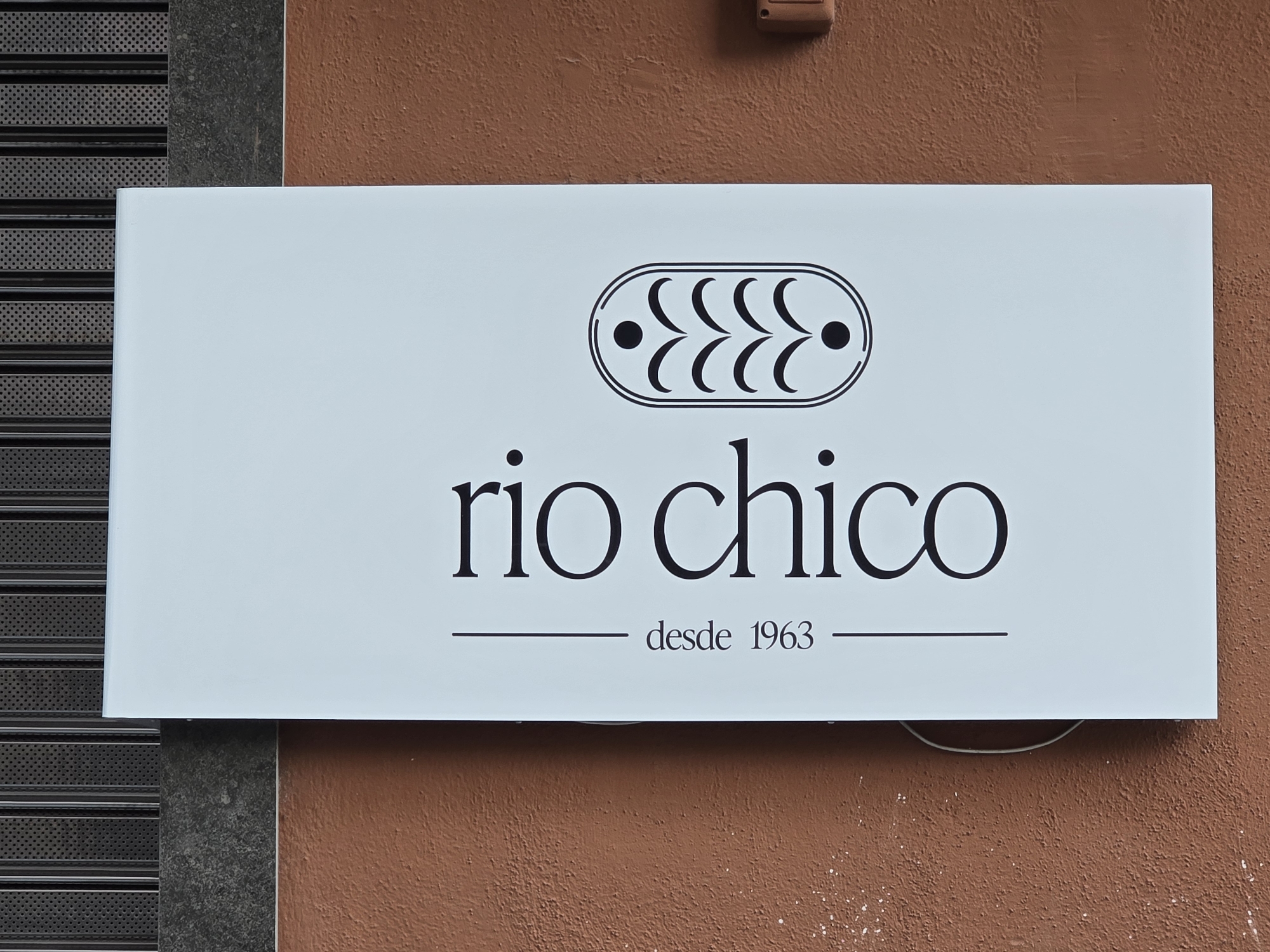 A horizontal white sign is affixed to a textured, terracotta-colored wall. To the left of the sign is a dark gray, perforated metal shutter. The sign displays the name "rio chico" in large, black, serif font. Above the name is a black, oval-shaped logo containing a series of stylized crescent shapes and two black dots. Below the name, in smaller black text, is "desde 1963," with thin horizontal lines to the left and right of this text. The sign appears to be an exterior storefront sign. The image appears to be taken in Jaén, Spain.