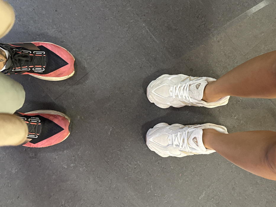 A top-down view captures the lower legs and footwear of two individuals standing on a dark gray, smooth floor. In the foreground, a person wears a pair of light-colored, off-white New Balance 2002R sneakers, with their bare, tanned lower legs visible. Positioned further back, another person wears black and pink athletic shoes, identified as Brooks running shoes by visible branding on the tongue, with part of their light-colored trousers visible and a small tattoo on the inner ankle of their right leg. Both individuals appear stationary. The scene is located in the city of Bangkok, Thailand.