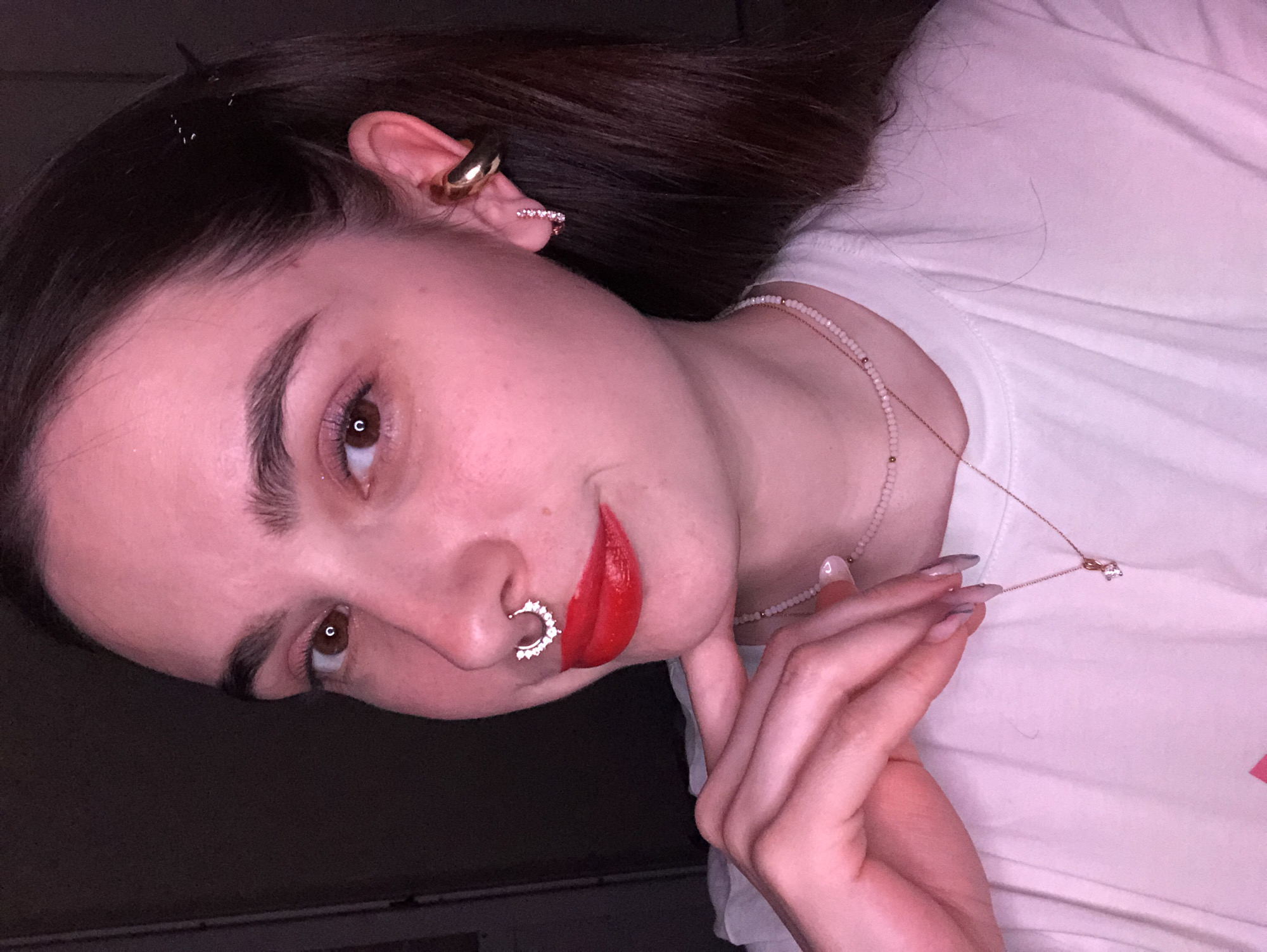 A woman is photographed at close range, with the camera positioned at a slightly high angle. She has dark hair, brown eyes, and fair skin. She is wearing red lipstick and has a silver septum piercing in her nose, as well as multiple ear piercings, including a gold cuff. She is wearing two necklaces. She is wearing a white t-shirt. The background is dark and indistinct. There is no evidence to suggest the location is Noviy, Russia.