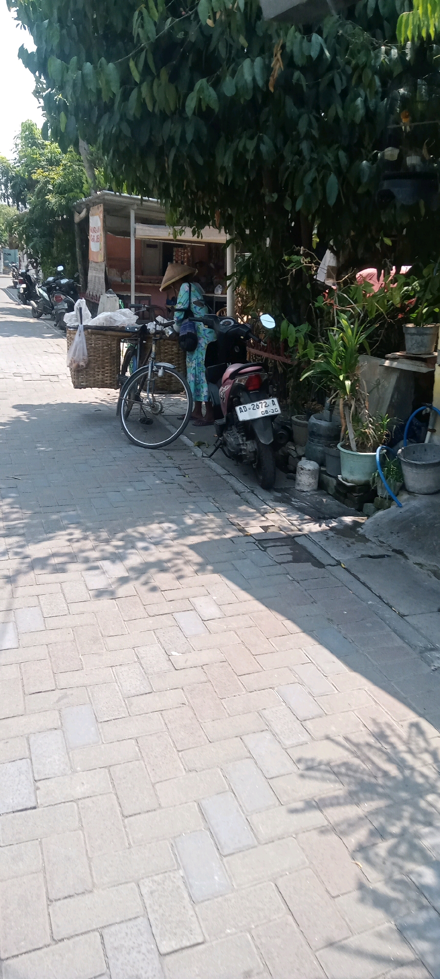 An outdoor scene in Surakarta, Indonesia shows a paved walkway lined with trees. A person wearing a conical hat and a long, patterned dress stands next to a bicycle laden with a woven basket and bags. A red scooter is parked to the right of the bicycle. The background features a small shop with a thatched roof and various potted plants. Sunlight casts shadows on the walkway.