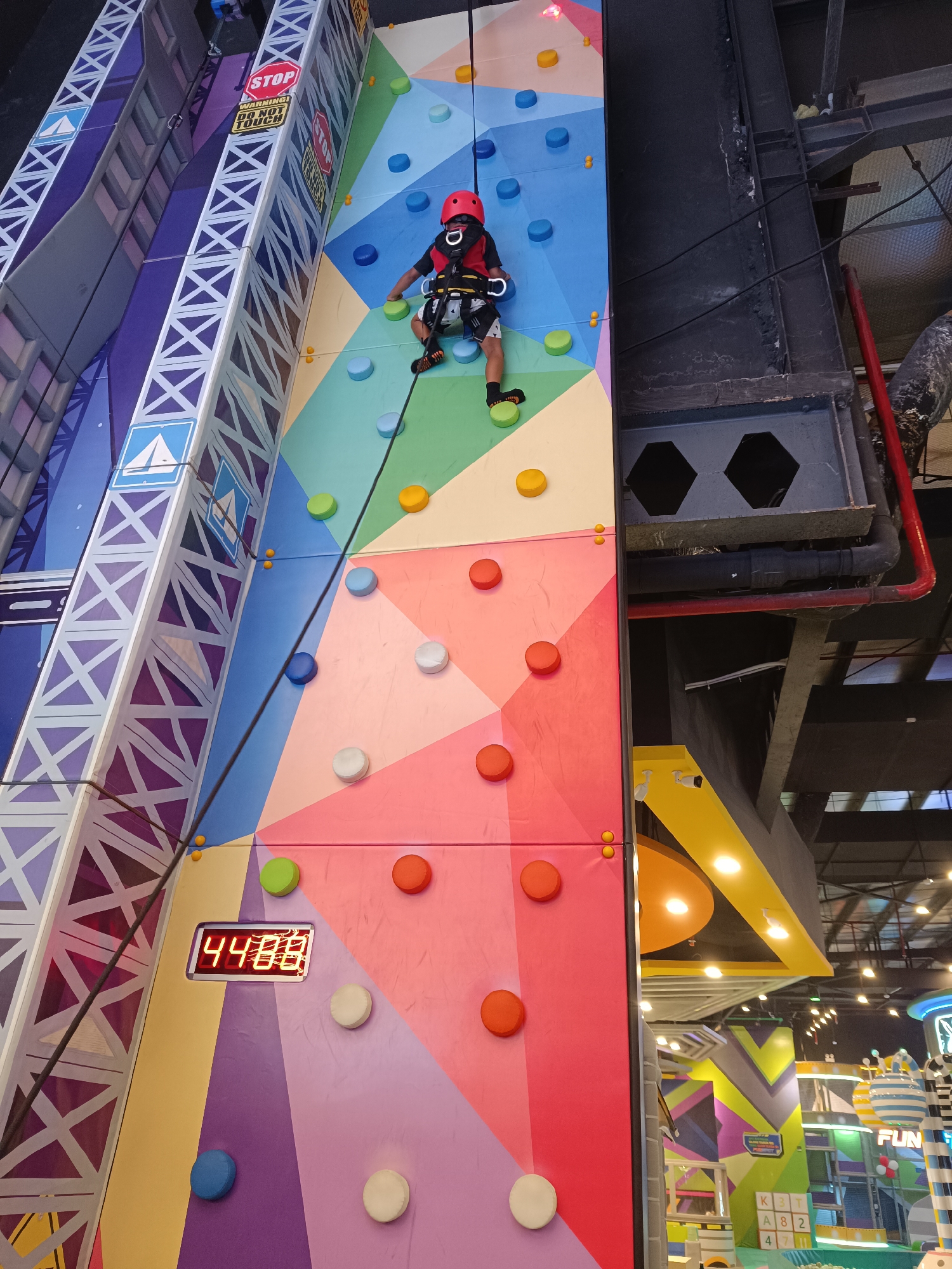 The image shows a young person wearing a red helmet and climbing gear on a colorful indoor climbing wall. The wall features various colored holds and geometric patterns. To the left of the climbing wall is a large, tall structure designed to look like a building facade with purple and white geometric patterns. A red digital timer displaying "44:08" is visible on the climbing wall. The background shows the interior of an entertainment venue with industrial-looking structures, exposed pipes, and lighting. The scene is located in Sorong, Indonesia.
