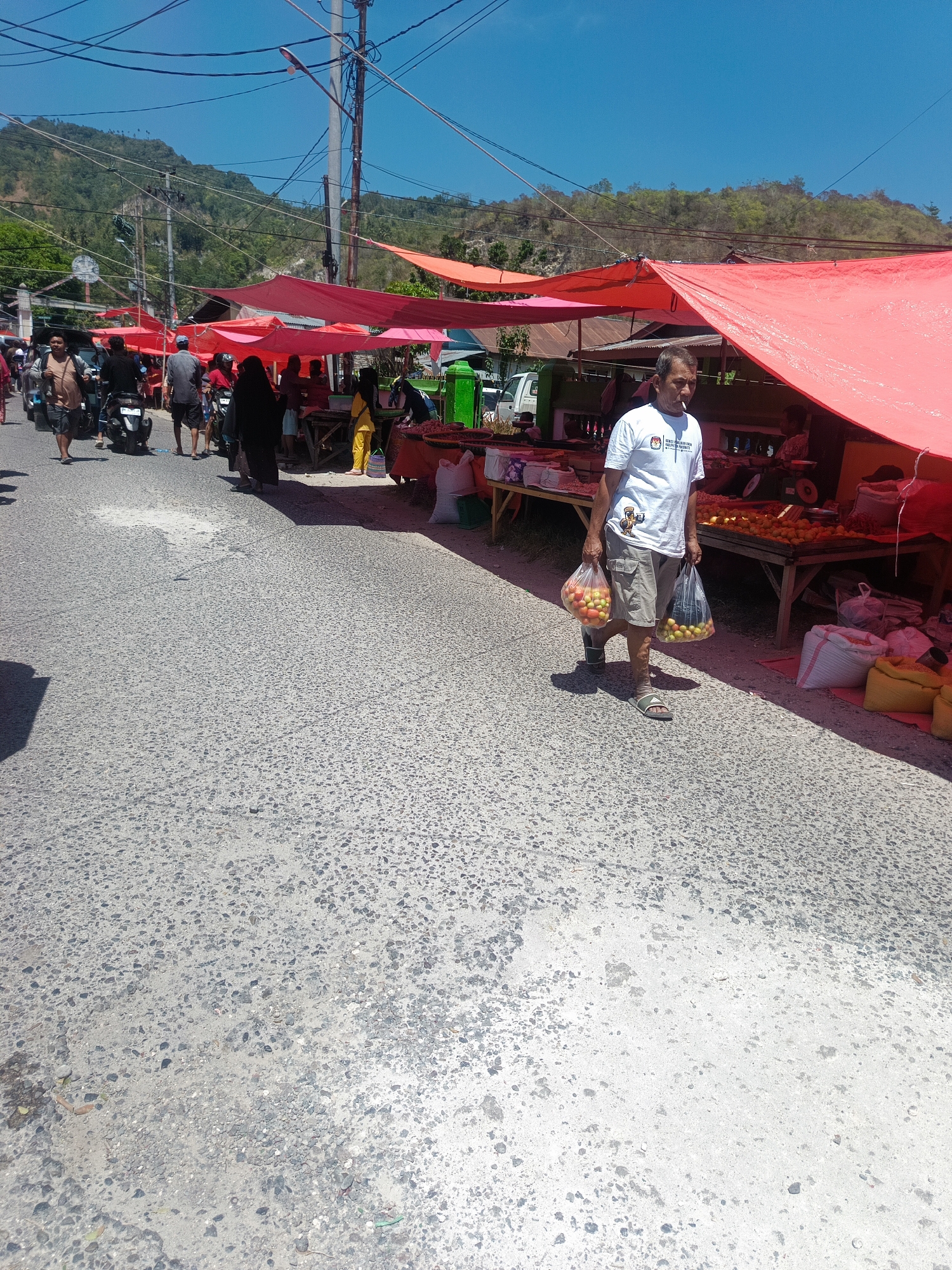A street market is set up along a paved road under a clear blue sky. Several stalls are covered by vibrant red tarpaulins, creating shade. People are seen browsing and shopping. In the foreground, a man wearing a white t-shirt and shorts walks carrying two plastic bags filled with produce, possibly tomatoes and oranges. He is smoking a cigarette. Behind him, a motorcycle is parked, and other people are visible walking or standing near the stalls. The background shows a sloping, green hillside and telephone poles with numerous wires. The overall scene depicts a lively daytime market activity in a tropical or subtropical urban environment.