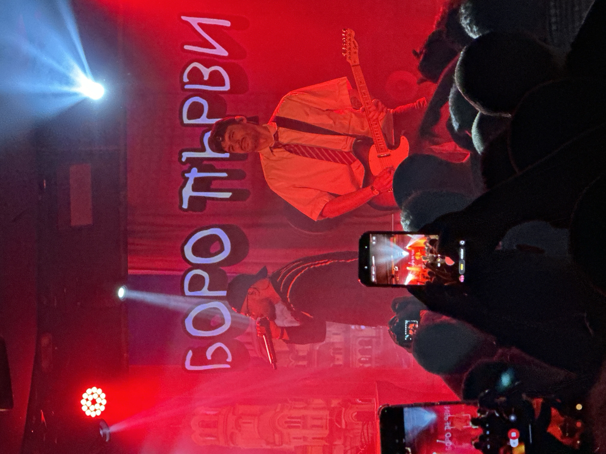 A concert stage at a venue in Sofia, Bulgaria. Two male performers are visible: one is playing a guitar and wearing a short-sleeved yellow shirt with a striped tie; the other, wearing a hat, is singing into a microphone. A backdrop behind them has lettering in what appears to be Cyrillic script. Stage lighting creates a red hue throughout the scene, with concentrated beams of light shining on the performers. The scene is viewed from the perspective of someone in the audience, as evidenced by the visible smartphones recording the performance.