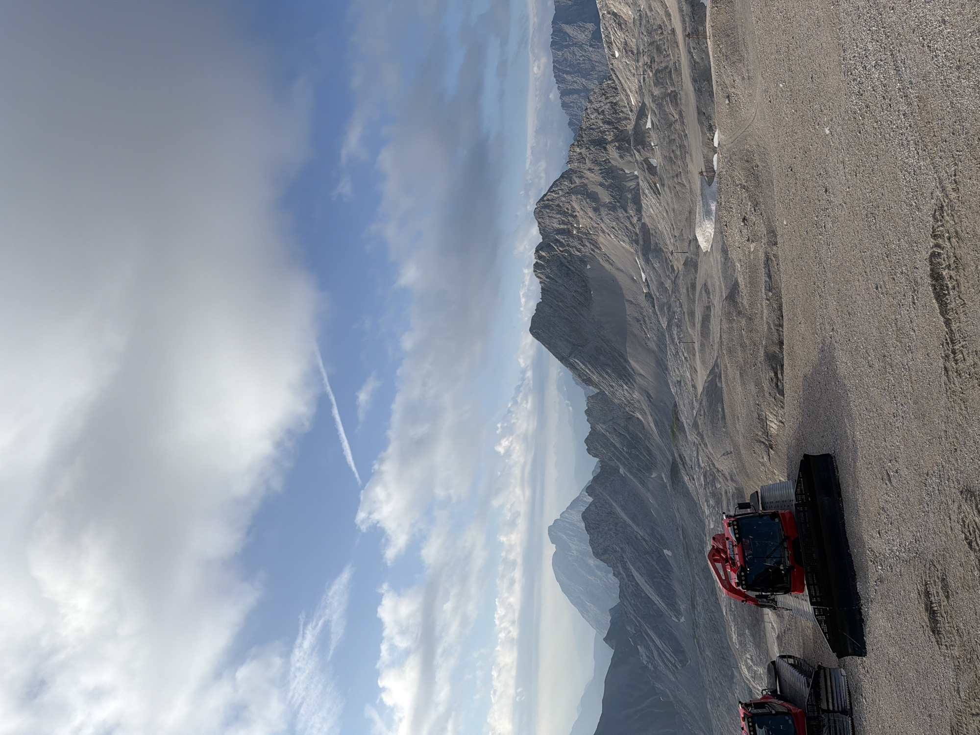 The image depicts a panoramic view of a mountainous landscape in Garmisch-Partenkirchen, Germany, likely at a high altitude, possibly on the Zugspitze. In the foreground, two red snowcats are parked on a gravelly, rocky terrain. The snowcats are heavy-duty vehicles with tracks, equipped with plows at the front. The larger snowcat in the center is more prominently visible, showcasing its red cab and mechanical arm.
The midground features a vast expanse of rocky, barren slopes that are characteristic of alpine environments. Sparse patches of snow are visible in crevices and depressions on the mountainsides. Several ski lift towers or similar infrastructure are scattered across the landscape, suggesting the area is used for winter sports or mountaineering.
In the background, a range of majestic, rugged mountains dominates the horizon. The peaks are sharp and rocky, with some showing traces of snow. The atmosphere is clear, with a hazy, muted quality in the distant peaks, indicating depth and distance. The sky above is a blend of blue and white, with scattered clouds that cast soft shadows on the mountains. A faint contrail from an aircraft is visible in the upper sky, cutting across the blue. The overall scene conveys a sense of vastness, ruggedness, and the raw beauty of the alpine wilderness.