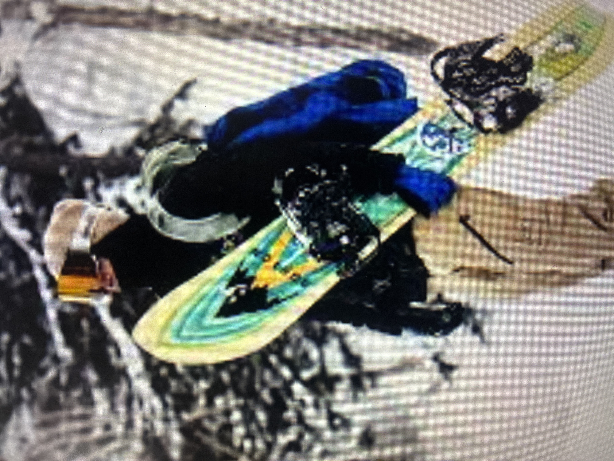 The image depicts a snowboarder in mid-air against a blurred, snowy background. The subject is wearing winter sports attire, including a black jacket or top, a vibrant blue layer (possibly a jacket or hoodie), and light brown or beige pants. They are also wearing goggles with an orange or yellow-tinted lens.
The snowboard is a prominent object, featuring a light green or yellow base color adorned with abstract, jagged patterns in shades of green, blue, and yellow. Black bindings are attached to the board. The dynamic pose of the snowboarder and the angle of the board suggest they are performing a jump or trick.
The scene is an outdoor, snow-covered environment, likely a mountain or a ski slope, indicating cold, winter conditions. The lighting suggests it is daytime. The background is blurred, emphasizing the movement and focus on the snowboarder. While the provided context specifies Asenovgrad, Bulgaria, the visual cues in the image itself are generic to a snowy mountain landscape and do not provide specific geographical confirmation.
Visible text is scarce and indistinct due to the image quality. A dark logo, possibly a Nike swoosh, can be faintly discerned on the light brown pants. A circular emblem with unidentifiable letters or symbols is also present on the snowboard near the front binding. The image has a grainy texture, possibly from being a photograph of a screen or an older capture.