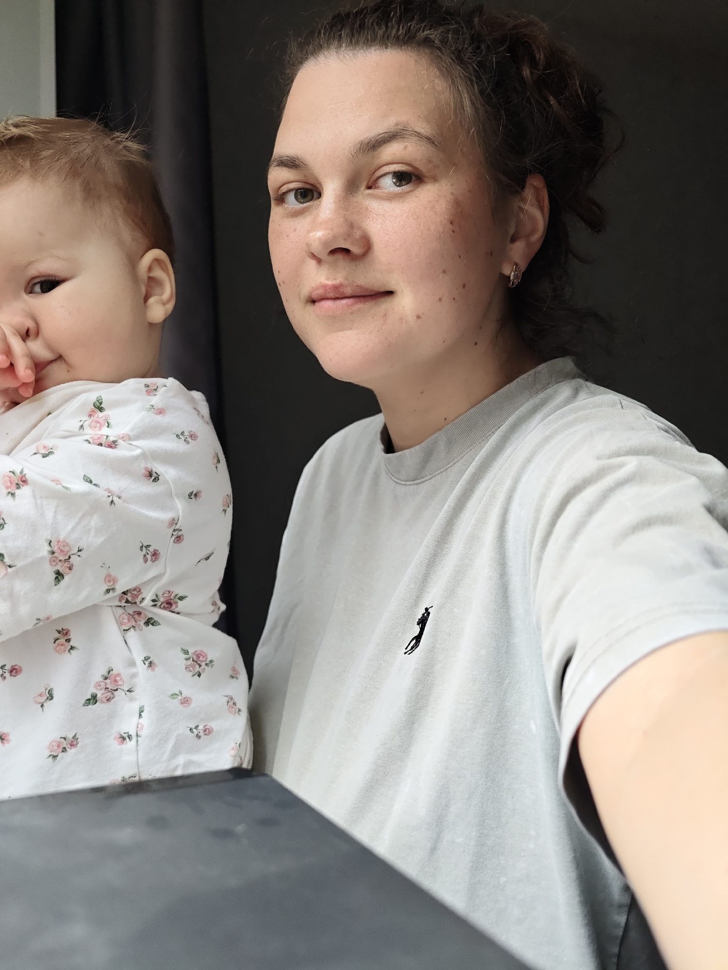 A woman holds a baby in front of her, both facing the camera. The woman has light skin, brown hair pulled back, freckles, and wears a grey t-shirt with a small black logo. The baby has fair skin, light brown hair, and wears a white floral-patterned shirt. The background appears to be a room with dark curtains or a dark wall. No other details suggest the location of Chaikovsky, Russia.