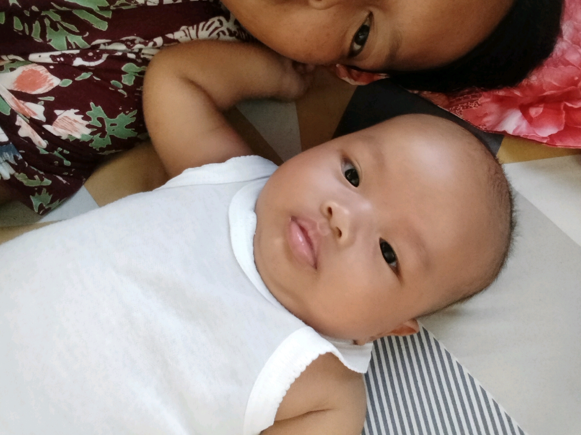 This is an overhead, eye-level medium shot of a baby lying on its back, looking towards the camera. The baby is wearing a white sleeveless shirt. To the left of the baby, a woman's upper body is visible, with her face close to the baby's head. She is wearing a maroon and green patterned shirt. Both individuals appear to be on a bed with a striped comforter. The setting is indoors, likely a bedroom. There is no specific information to pinpoint the location within Gempolan, Indonesia.