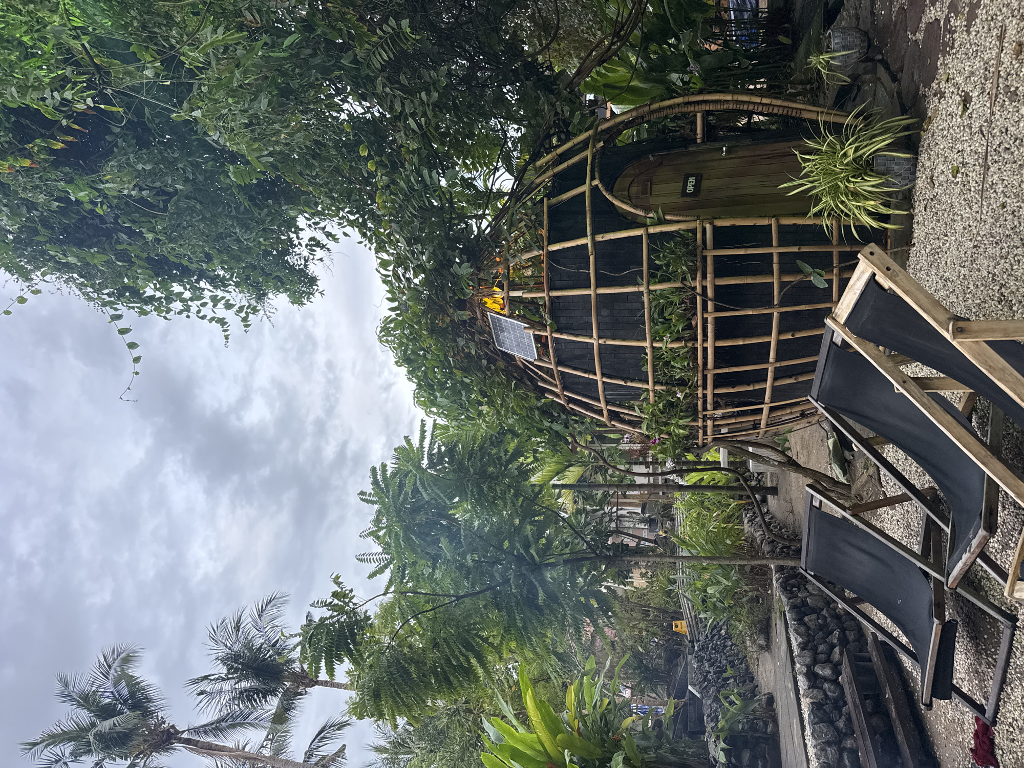 The image depicts a tranquil outdoor setting in Baan Sri Thanu, Thailand. A circular structure, possibly a small shop or dwelling, is framed by a bamboo lattice and surrounded by lush tropical foliage. The door bears a sign reading "OPEN". A solar panel is visible on the structure. In the foreground, two black lounge chairs suggest a relaxed atmosphere. The ground is covered with small gravel, and a low stone wall adds to the naturalistic feel. Palm trees and other tropical plants are prevalent, creating a verdant environment. The sky is overcast, indicating a possibly rainy or cloudy day.