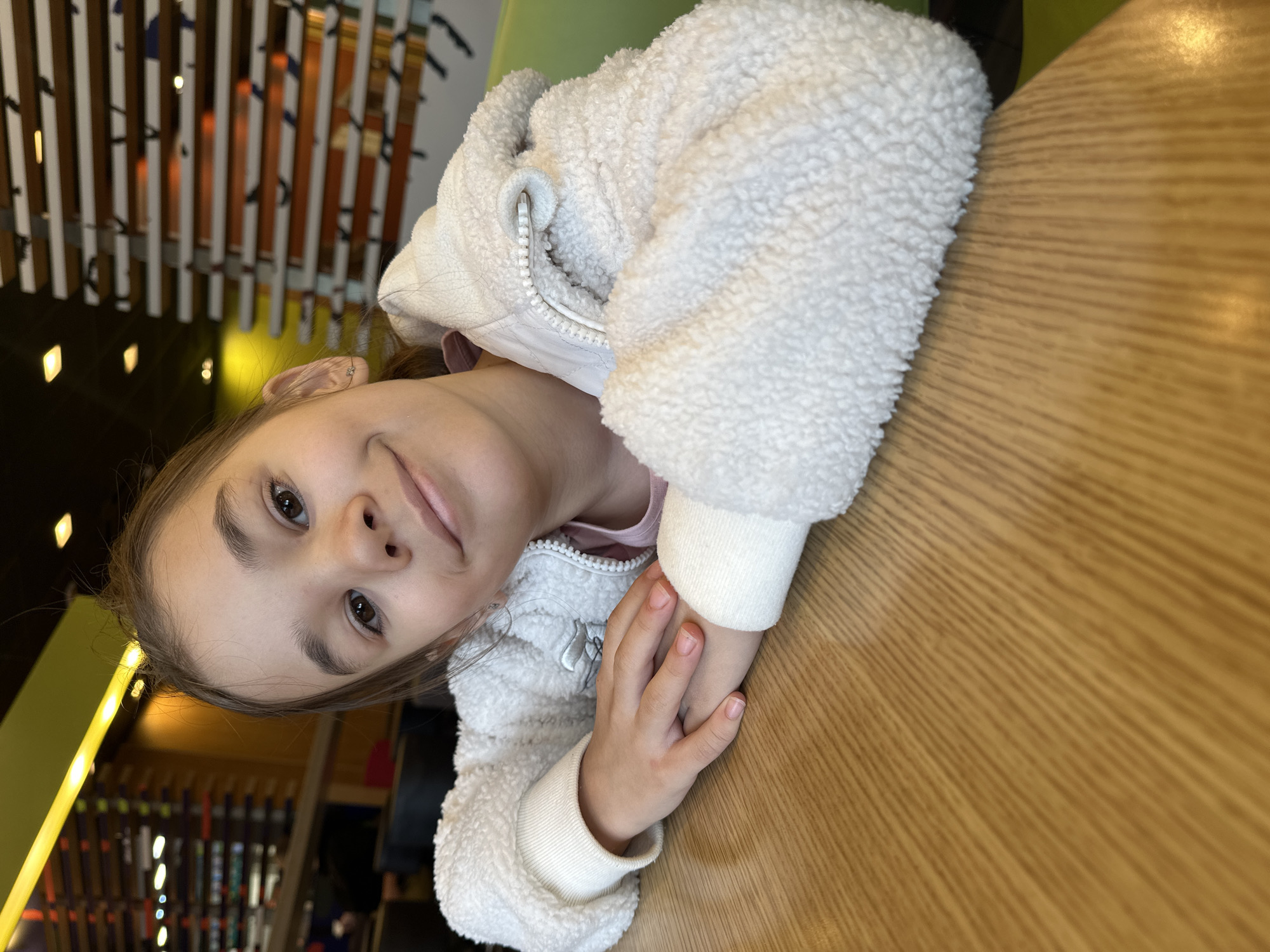 A young girl wearing a white sherpa jacket sits at a wooden table. She has light brown hair pulled back and is looking directly at the camera with a slight smile. Her hands are resting on the table in front of her.
In the background, out of focus, there are vertical elements possibly depicting a building or structure. Based on the presence of skis, it is likely the photo was taken in a building with skiing equipment in the background. Given the location prompt is Izhevsk, Russia, this is likely a ski resort, shop or restaurant in that city.