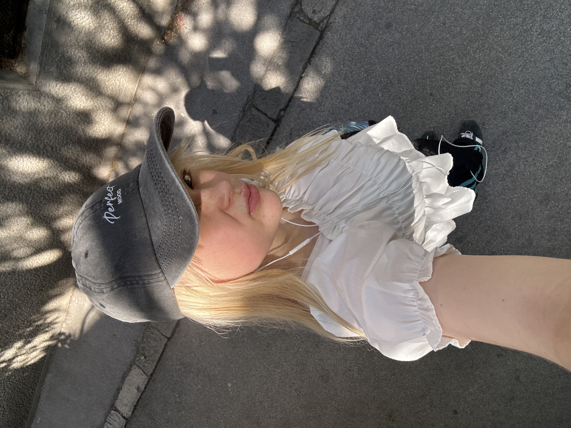 A young woman with blonde hair and fair skin is shown from a high angle, looking upwards. She is wearing a grey baseball cap with white text that reads "Perfect Mood" and a white ruffled blouse. White earbuds are visible in her ears. She is standing on a paved surface, possibly a sidewalk, with dappled sunlight casting shadows from overhead foliage. In the background, her feet are visible, wearing black roller skates with teal accents. The setting appears to be outdoors on a sunny day, likely in a park or urban area. The lighting suggests it is daytime.