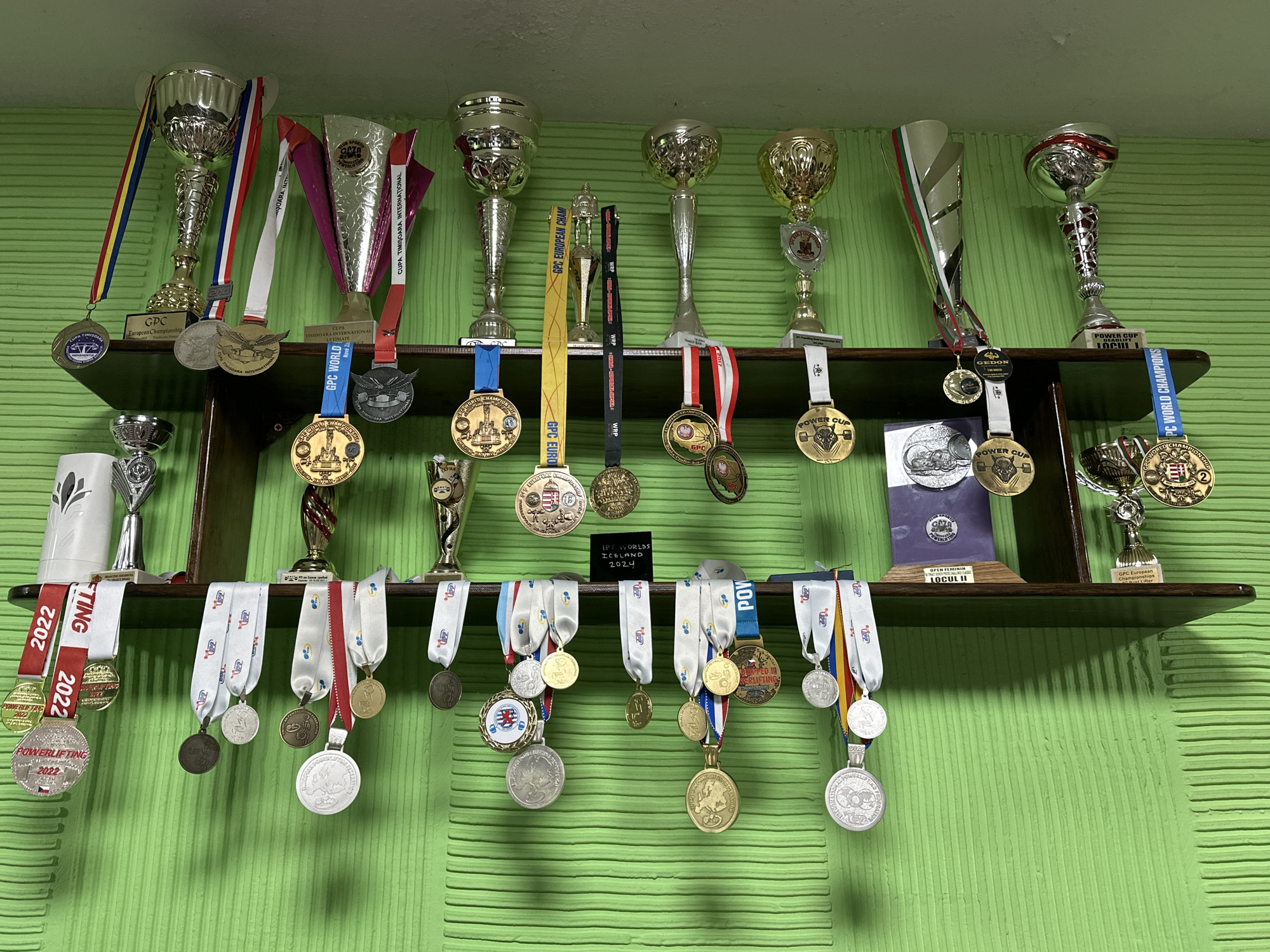 The image shows a two-tiered dark brown wooden shelf mounted on a textured, light green wall. The shelf is laden with a collection of trophies and medals. The trophies, made of silver and gold-colored metal, vary in size and design and are arranged along the top and bottom shelves. Medals hang from the front edges of both shelves, suspended by ribbons in a variety of colors and patterns. The medals themselves appear to be made of gold, silver, and bronze-colored metal. Some medals are marked with the year "2022" and the word "Powerlifting." In the middle of the bottom shelf, a small black sign with white lettering reads "IPF WORLDS ICELAND 2024". Additional text on the medals and trophies includes words and phrases like "European Championship", "GPC World", and "Power Cup".