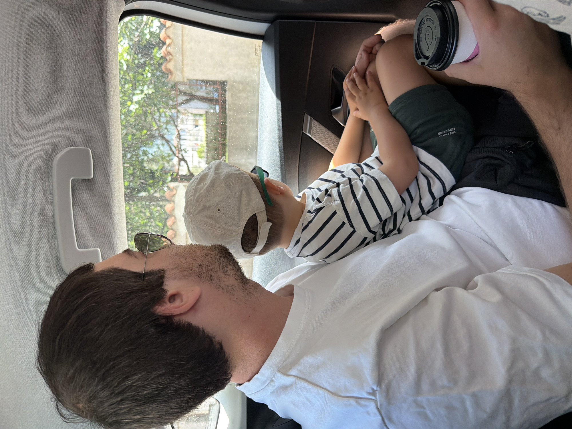 Inside a vehicle, a man and a child are seen. The man, with dark hair and sunglasses perched on his head, is wearing a white t-shirt. He is holding a white and pink disposable coffee cup with a black lid in his hand. A young child, possibly a boy, is resting on the man's lap or chest. The child wears a light-colored baseball cap, a white t-shirt with black horizontal stripes, and dark green shorts. His hands are clasped near his knees.
The setting appears to be the back seat of a car or similar vehicle, characterized by a light gray interior pillar/ceiling trim and a white grab handle. Through the window, a bright, sunny outdoor scene is visible, featuring green foliage from trees or bushes and parts of buildings with what appear to be terracotta-colored roofs or tiles, consistent with the provided location context of Budva, Montenegro. The overall impression is one of calm, with the child resting comfortably against the man, suggesting they are on a journey or simply relaxing together.
Notable details include the text "THE F_RC_ / EXPLORING" (partially obscured, likely "THE FORCE EXPLORING") on the child's dark green shorts. The bright light outside indicates it is daytime. No other clear text is legible on the coffee cup lid.