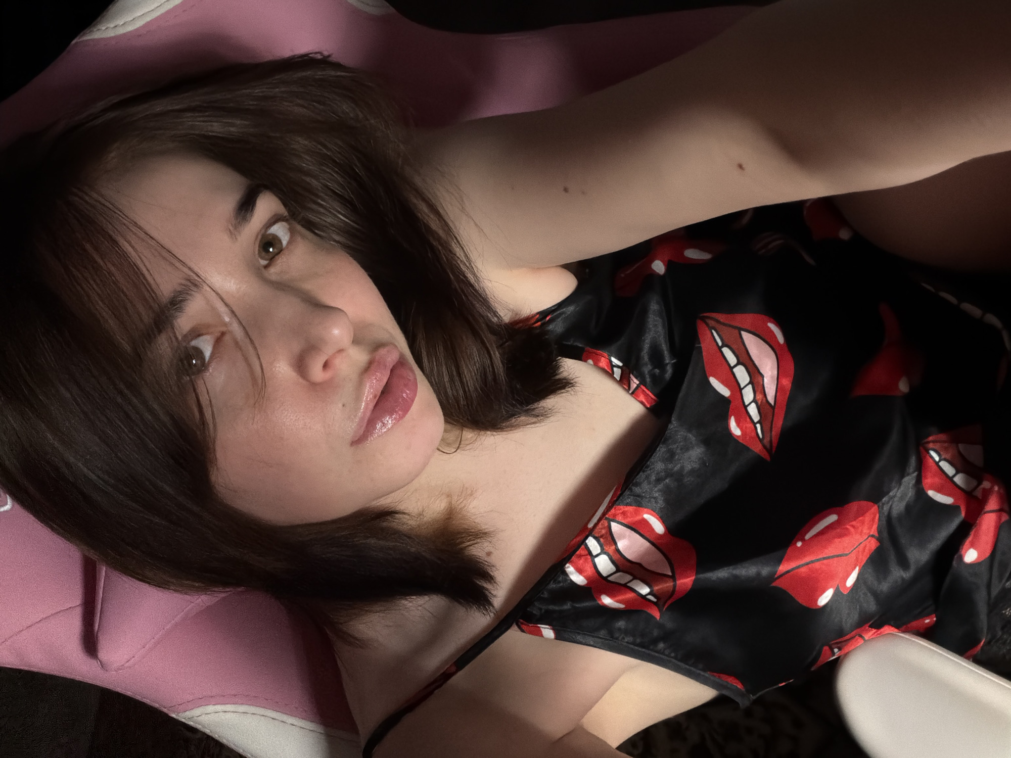 A close-up, high-angle shot shows a young woman with dark brown hair and light brown eyes, leaning back in a pink and white gaming chair. She is wearing a black satin camisole with a bold red and white lip print pattern. Her hair is styled in a short, choppy bob, and she is looking towards the camera with a slight pout. The lighting is dim, creating a soft glow on her skin and highlighting her glossy lips. The background is dark, with the pink of the chair being the most prominent color. The scene appears to be an indoor setting, possibly a bedroom or gaming room, with no identifiable landmarks or context to suggest a specific location within a city.