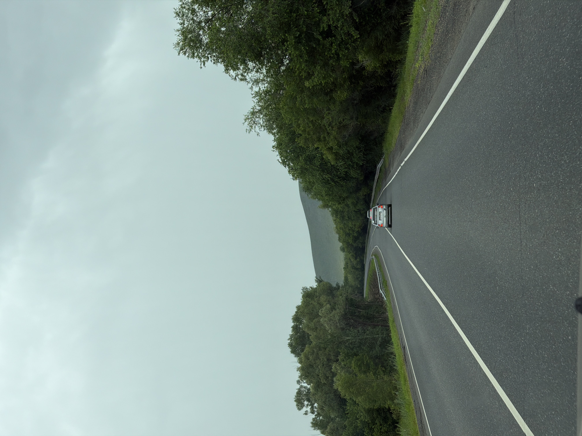 A vehicle, a silver sedan with a visible license plate, is traveling on a two-lane asphalt road in Верхнеозёрное, Russia. The road is marked with a solid white line on the left and a broken white line on the right, indicating a clear lane separation. The car is in the right lane, heading away from the viewer. The environment consists of lush green trees lining both sides of the road, with a gentle hill in the background. The sky is overcast and grey, suggesting a cloudy or possibly rainy day. The overall scene depicts a scenic drive through a rural or natural landscape.
