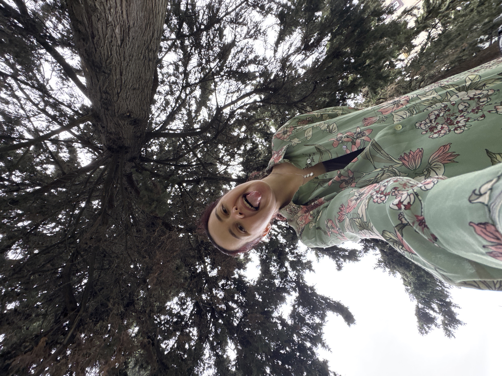A low-angle selfie depicts a woman with auburn hair and fair skin wearing a light green floral-patterned blouse. She is smiling with her tongue sticking out. A silver necklace is visible around her neck. Behind her is a large tree with dense, dark green foliage and a thick, gnarled trunk. The sky is overcast and white. The location is potentially in Sochi, Russia, based on visual characteristics of the tree.