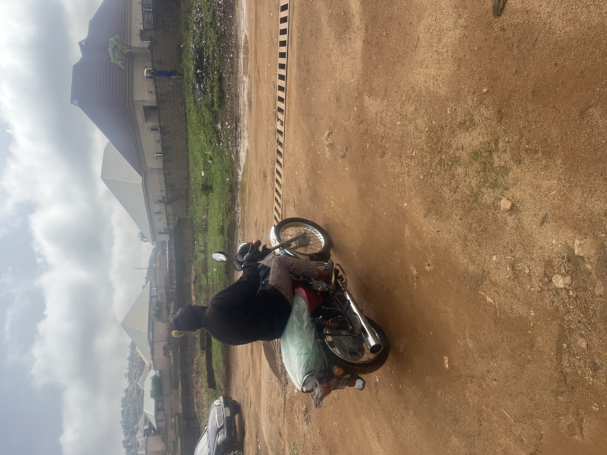 The image shows a person sitting on a red motorcycle on a dirt road. The person is wearing a black hooded sweatshirt and patterned pants. A large bag is strapped to the back of the motorcycle. A dark colored car is parked nearby on the left side of the road. The background includes buildings and vegetation. The location is in the city of Barakin Akawo, Nigeria. The ground is dirt. There is a black and white stripe barrier in the background. The sky is cloudy.