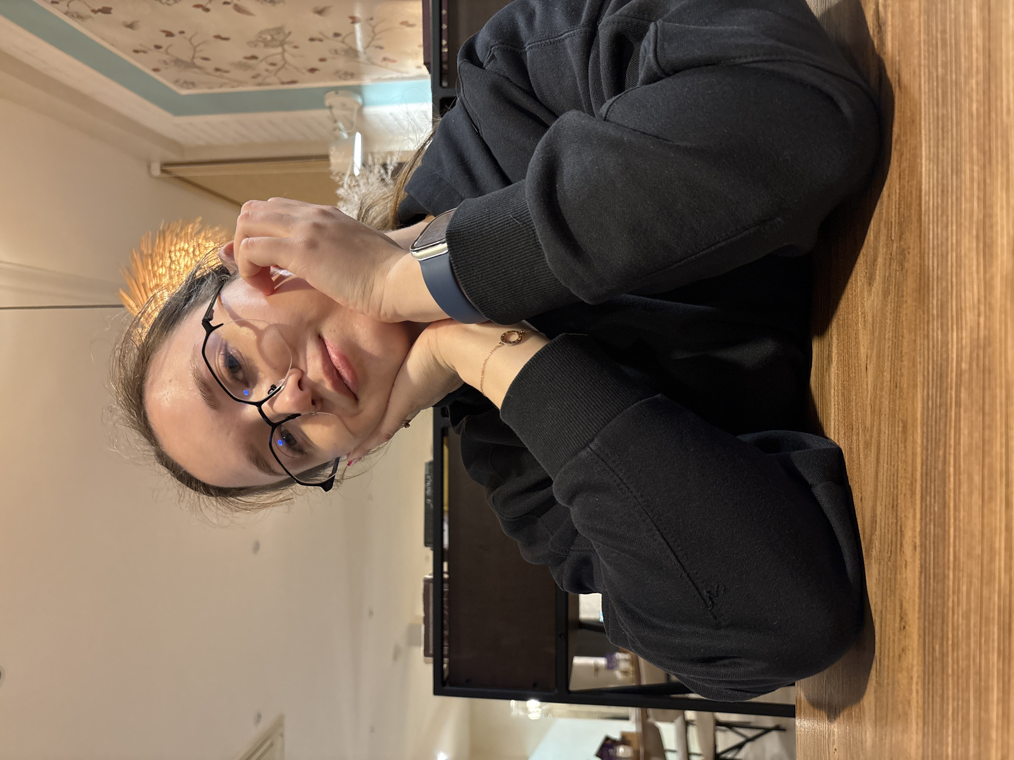 A person wearing glasses, a black hooded sweatshirt, and a golden straw crown is seated at a wooden table in what appears to be an indoor setting. The person's hands are resting on their face. The background is slightly blurred, suggesting an interior space with light-colored walls and decorative elements. The location in Izhevsk, Russia, cannot be verified from the image alone.